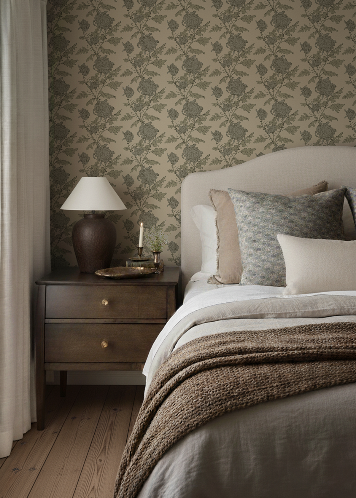 A cozy bedroom corner with a nightstand, table lamp, candle, floral decor, and neatly made bed with several pillows and a knitted blanket, set against patterned wallpaper.