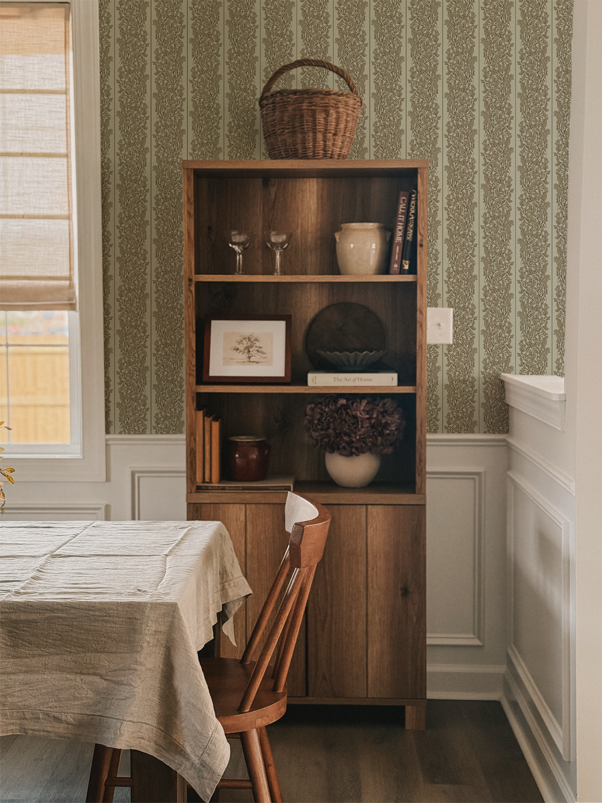 A wooden bookshelf with three shelves, decorated with various items including books, vases, and art, in a dining room with patterned wallpaper and a window.