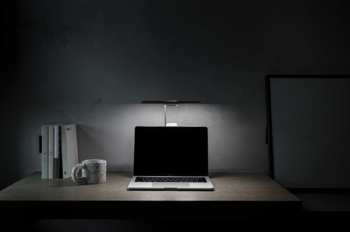 Desk with a laptop, a mug, and books under a lamp, against a gray wall.