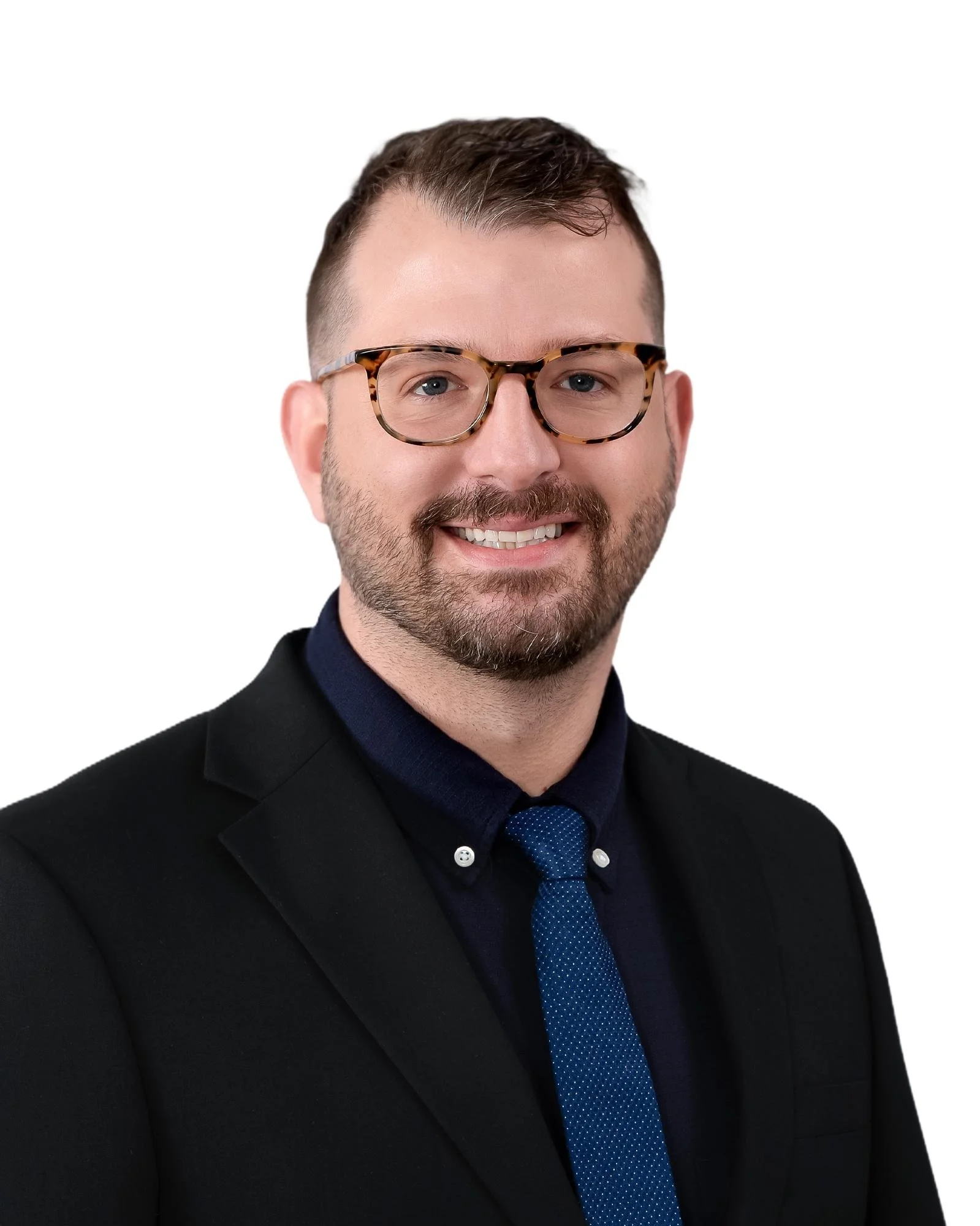 A headshot of a man with short brown hair, wearing tortoiseshell glasses, a dark suit, and a blue tie with small white polka dots, smiling against a plain white background.