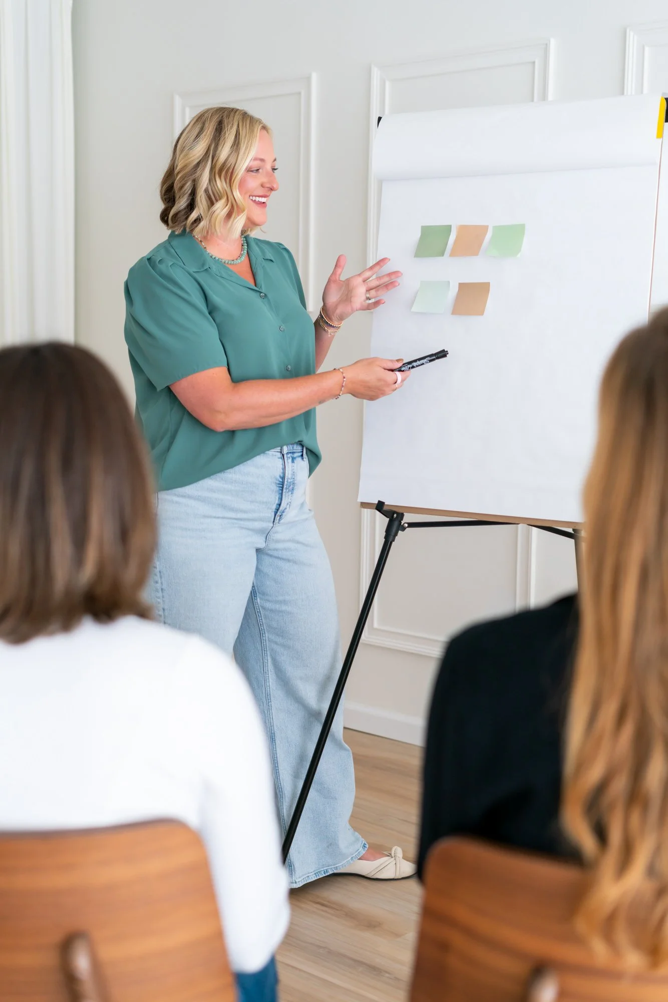 Woman giving a presentation to a small group, standing next to a whiteboard with sticky notes, in a bright room.