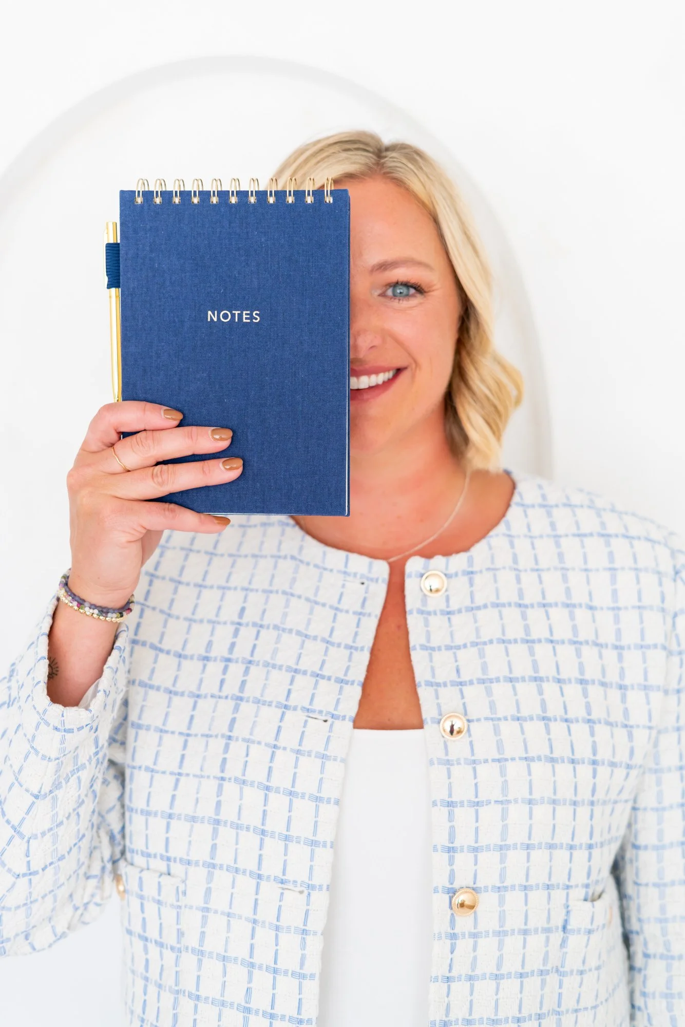 A smiling woman with blonde hair holding a blue spiral notebook labeled 'NOTES' in front of her face. She is wearing a white and blue checkered jacket with gold buttons and a white top underneath, accessorized with colorful bracelets and a silver necklace.