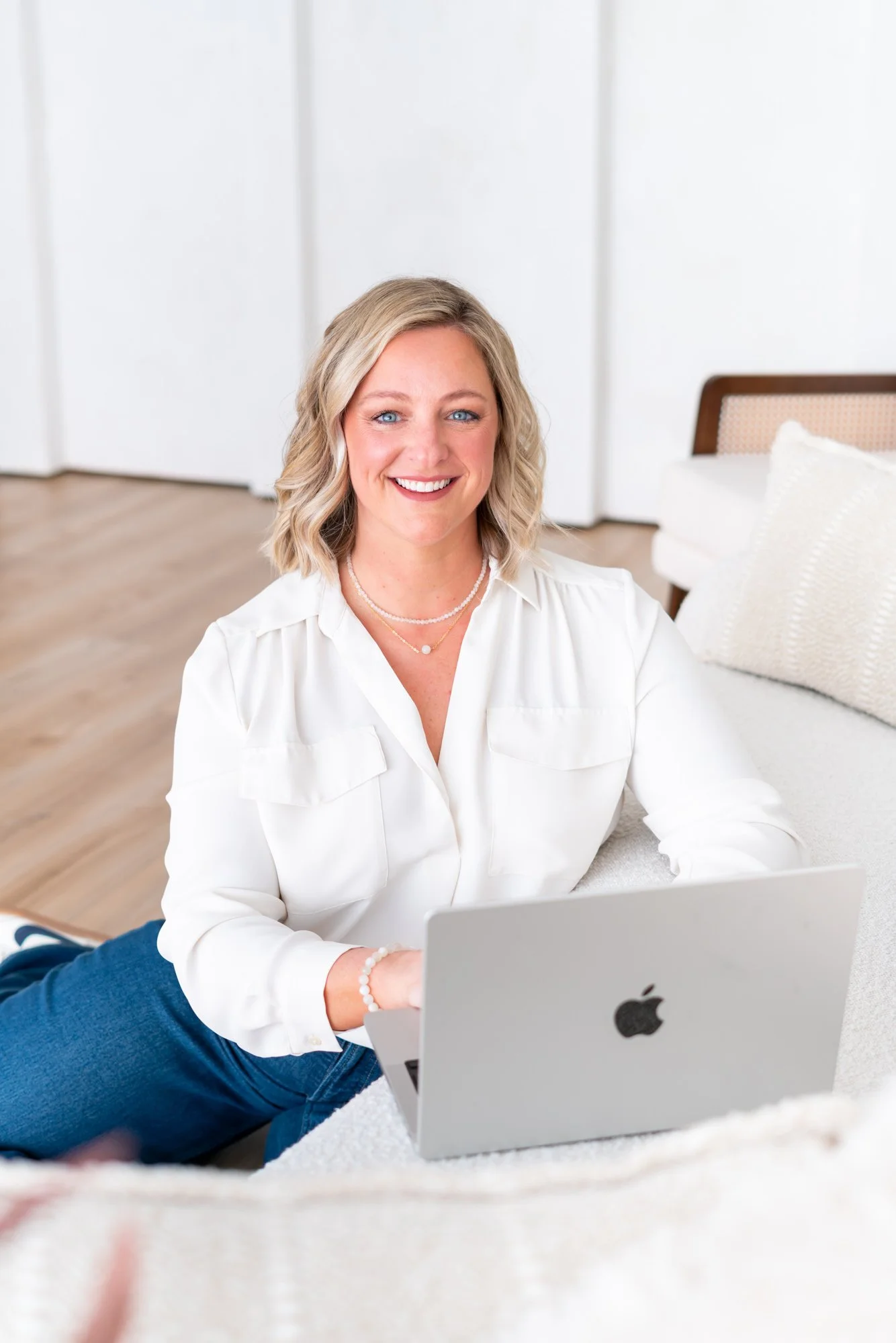 A woman with blonde hair and blue eyes smiling, sitting on the floor with a silver laptop in front of her. She is wearing a white shirt and jewelry, in a bright room with a bed and pillows in the background.