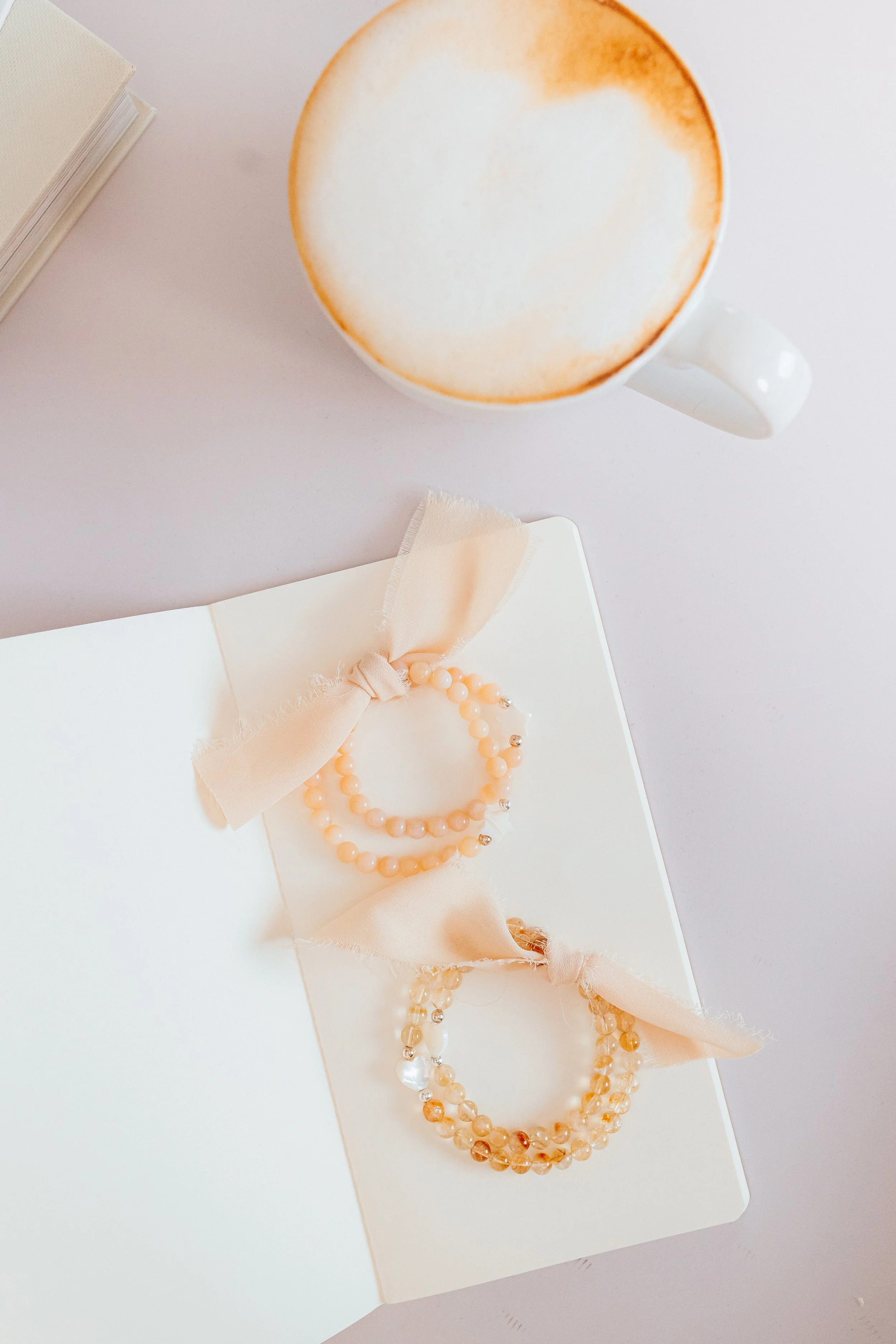 A white open jewelry box with pearl and peach-colored beaded necklaces, a peach-colored bow, and a beige fabric ribbon inside.