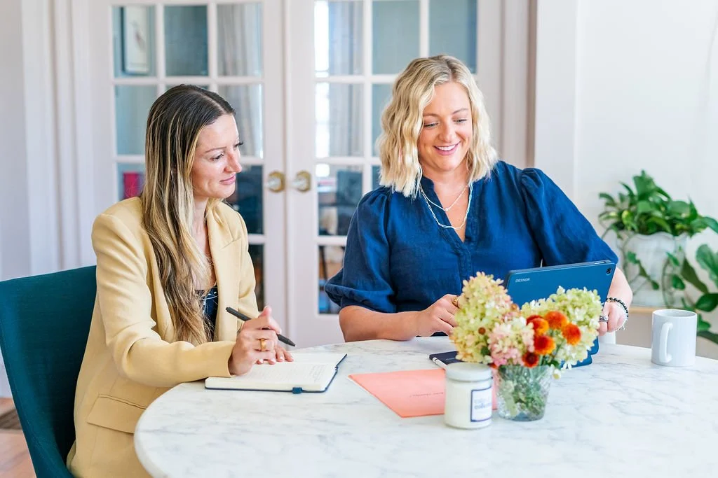 Two women sitting at a round table in a bright room, one with blonde hair looking at a tablet, the other with brown hair taking notes, with flowers and a mug on the table.