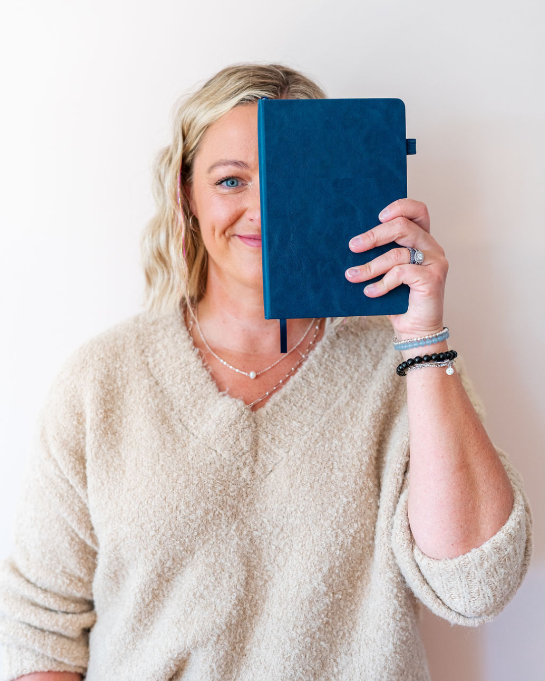 A woman with blonde hair, wearing a beige sweater, holding a blue notebook in front of her face and smiling.