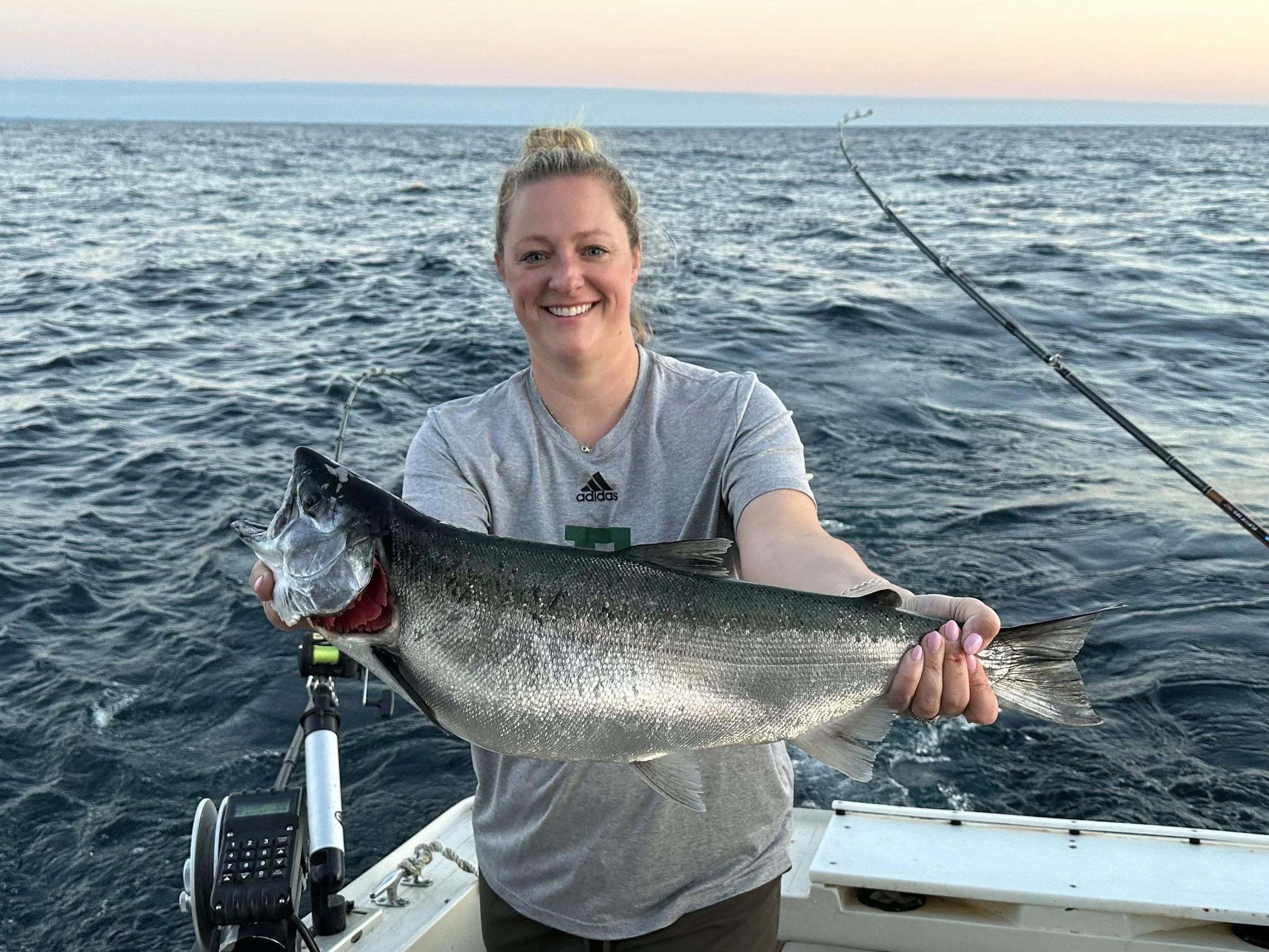 Woman smiling on a boat holding a large fish she caught while fishing in the ocean at sunset.