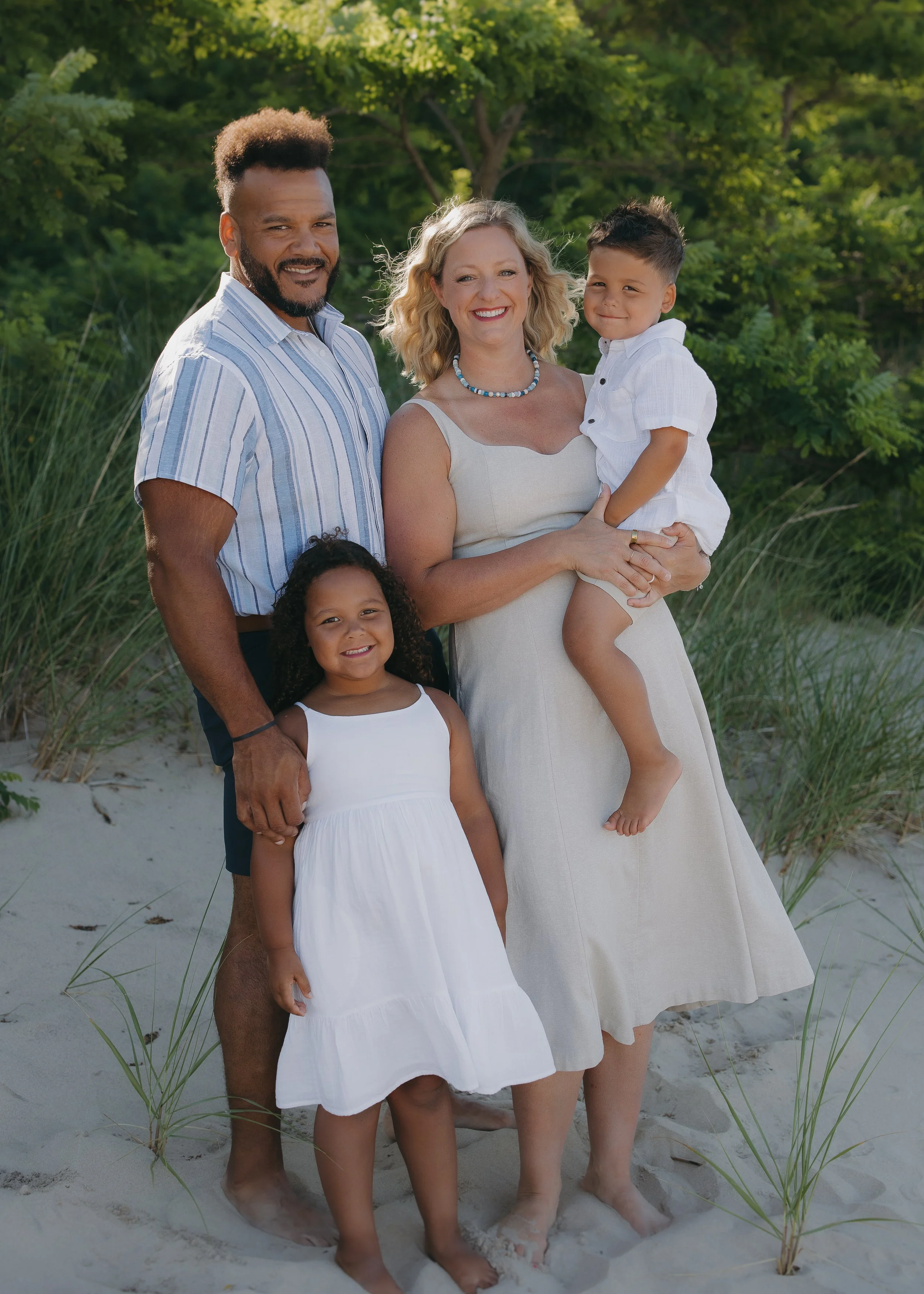 A happy multiracial family of five stands on a sandy beach with green trees in the background. The father, mother, and three children are smiling, dressed in light summer clothing, with the mother holding a young boy in her arms.