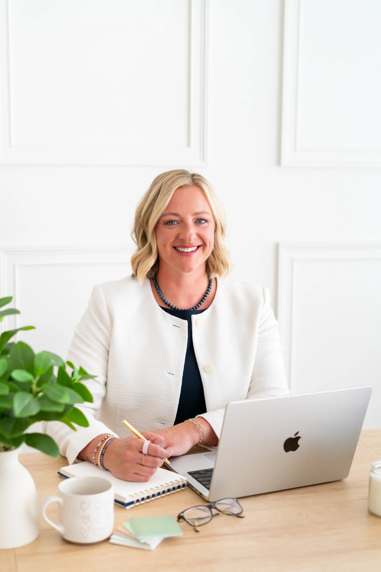 A woman with blonde hair sitting at a wooden desk, working on a silver laptop, surrounded by office supplies and plants.