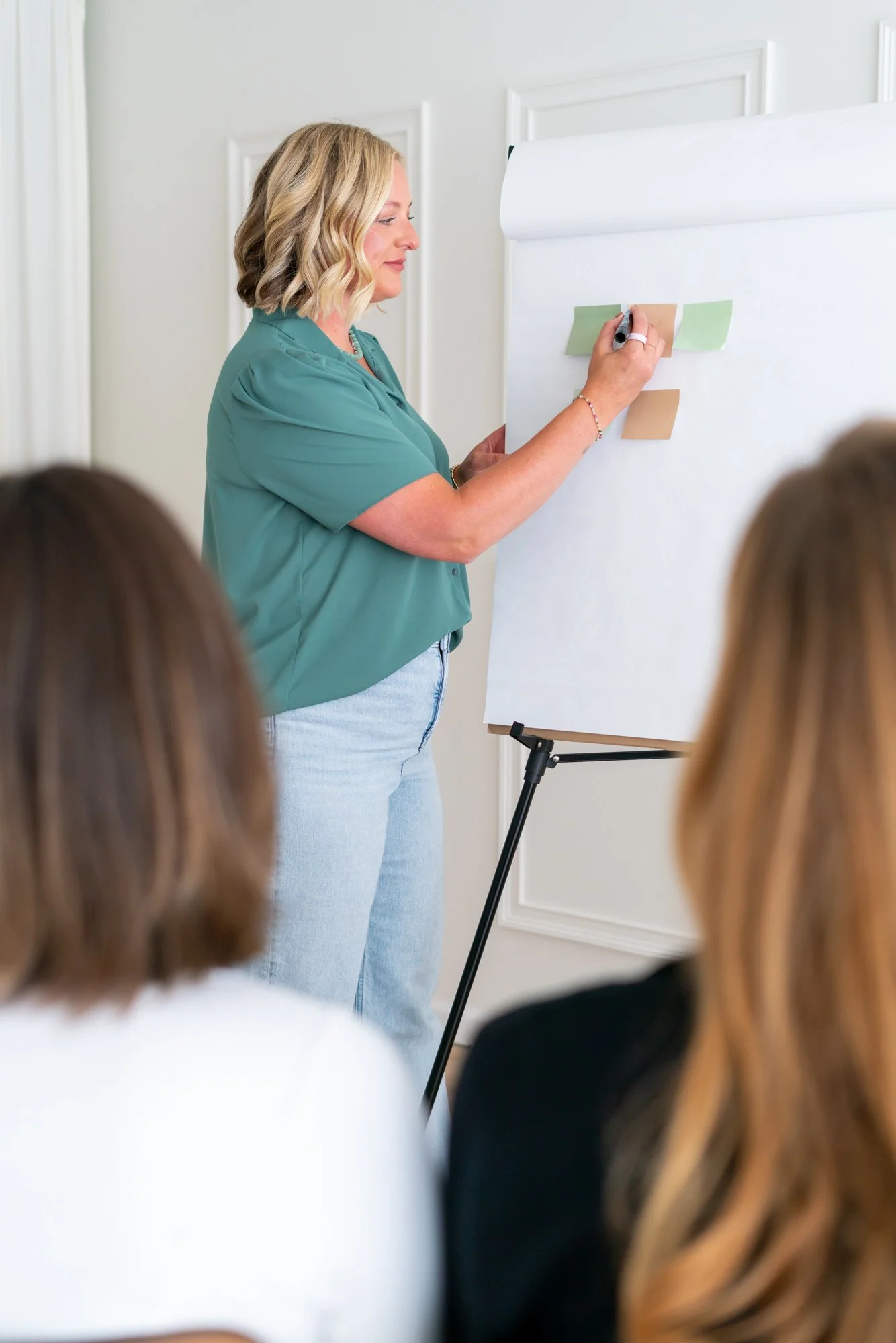 A woman with blonde hair in a green shirt is giving a presentation to a group of people, writing on a large white presentation board with note papers attached.