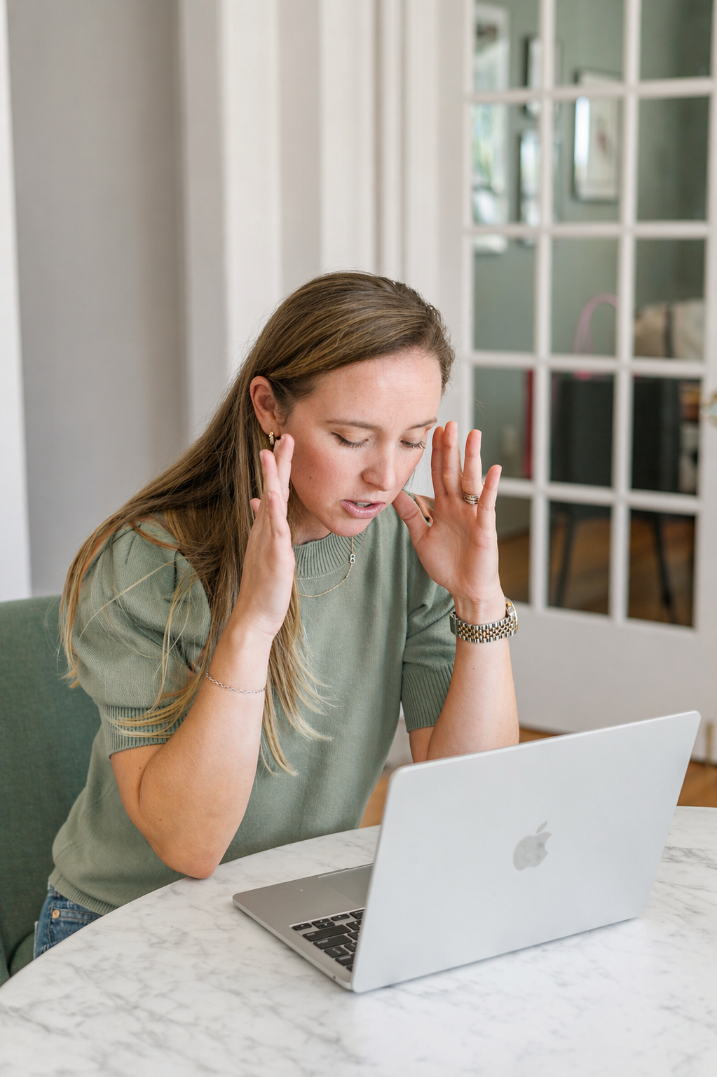 A woman sitting at a marble table with a silver MacBook laptop, holding her head with a stressed expression, wearing a green shirt, jewelry, and a watch, in a bright, modern room.