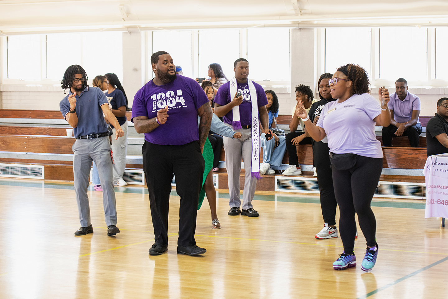 Group of people dancing and socializing in a gymnasium during an event, with some sitting on bleachers in the background.