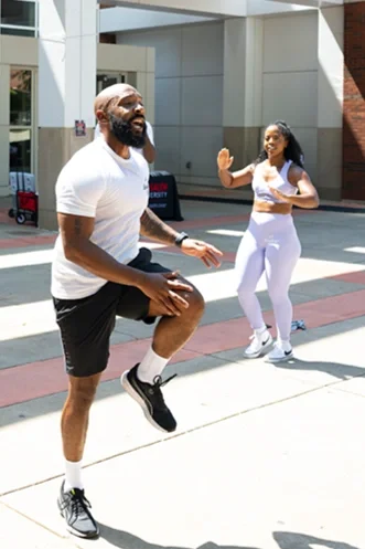 A man and woman exercising outdoors, with the man performing a knee lift and the woman giving a high-five.