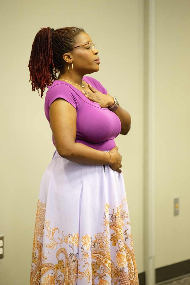 A woman with glasses and earrings, wearing a purple top and a long white skirt with orange and gold floral patterns, stands with one hand on her chest and the other on her stomach, appearing to be in a moment of reflection or prayer.