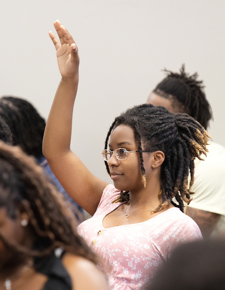 A young woman with glasses and curly dreadlocks raises her hand in a classroom or meeting room.