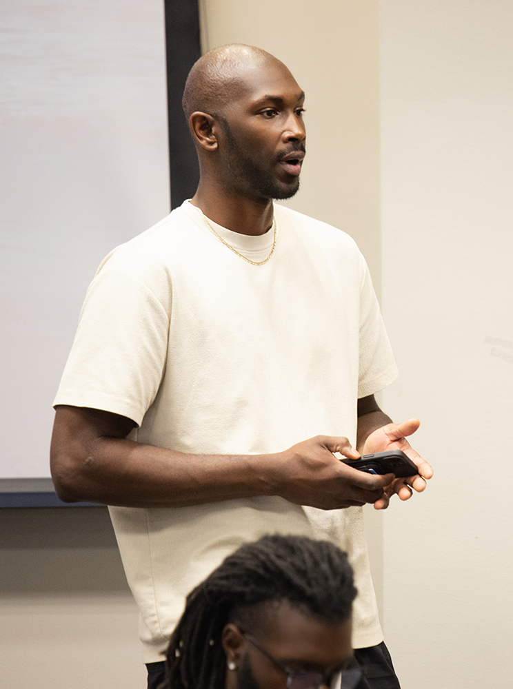 A young man with a shaved head and beard speaking in a classroom or conference room, holding a phone, with a woman with glasses and braided hair sitting in front of him.