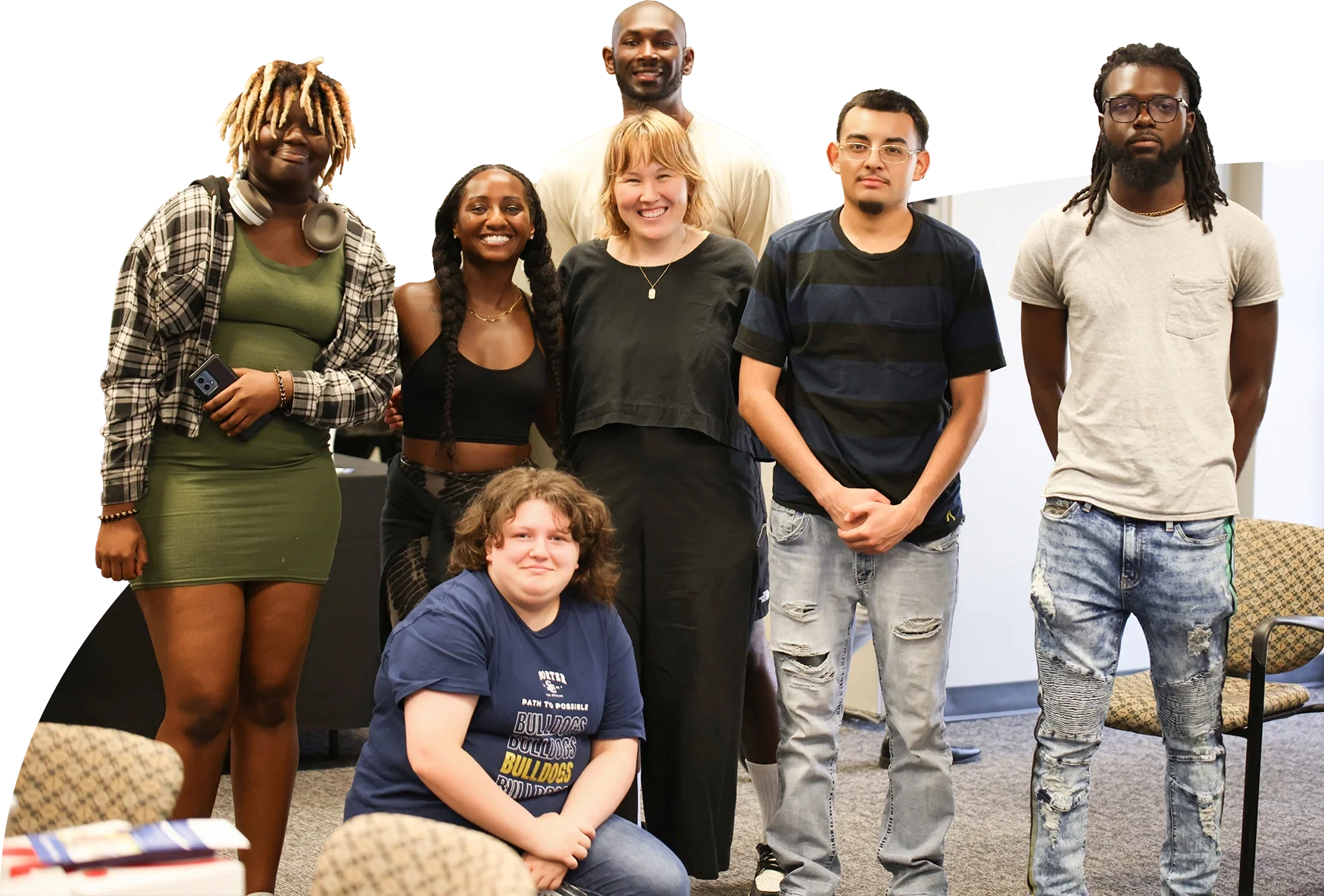 Group of seven diverse people, including children and adults, smiling and posing indoors, with one seated on the floor and others standing, in a casual setting.