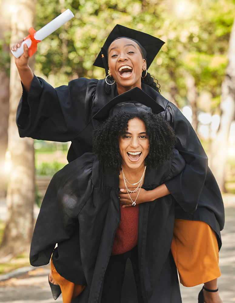 Two women in graduation caps and gowns celebrating outdoors, one giving a piggyback ride to the other, both smiling and joyful.
