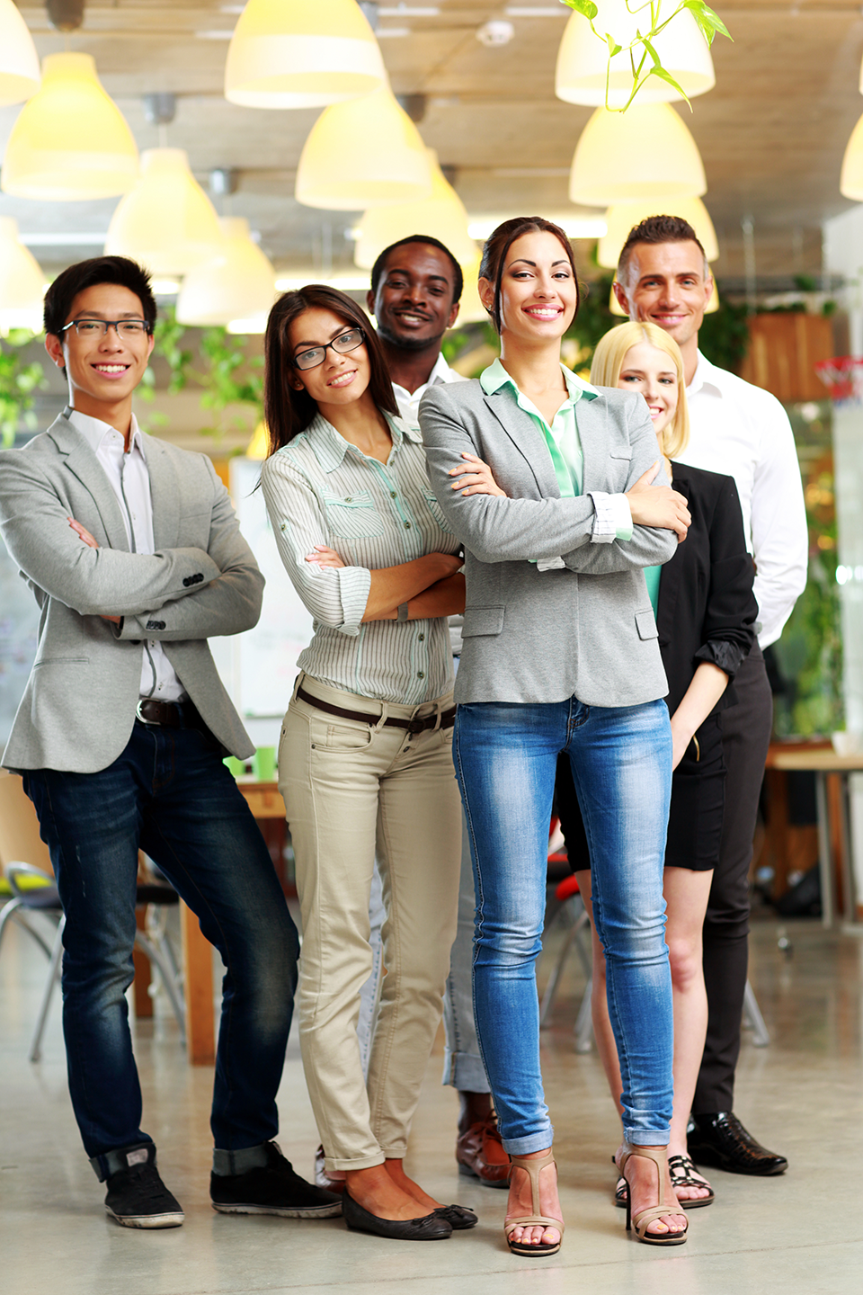 Group of six diverse professionals standing together in an office with warm lighting and green plants, smiling confidently.
