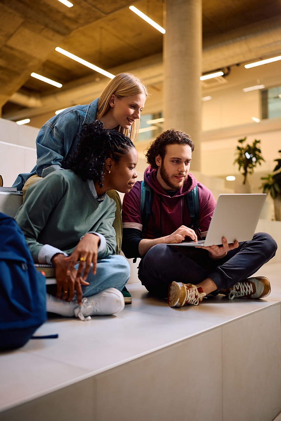 Three young adults sitting on steps in an indoor space, looking at a laptop together, with one person standing behind them.