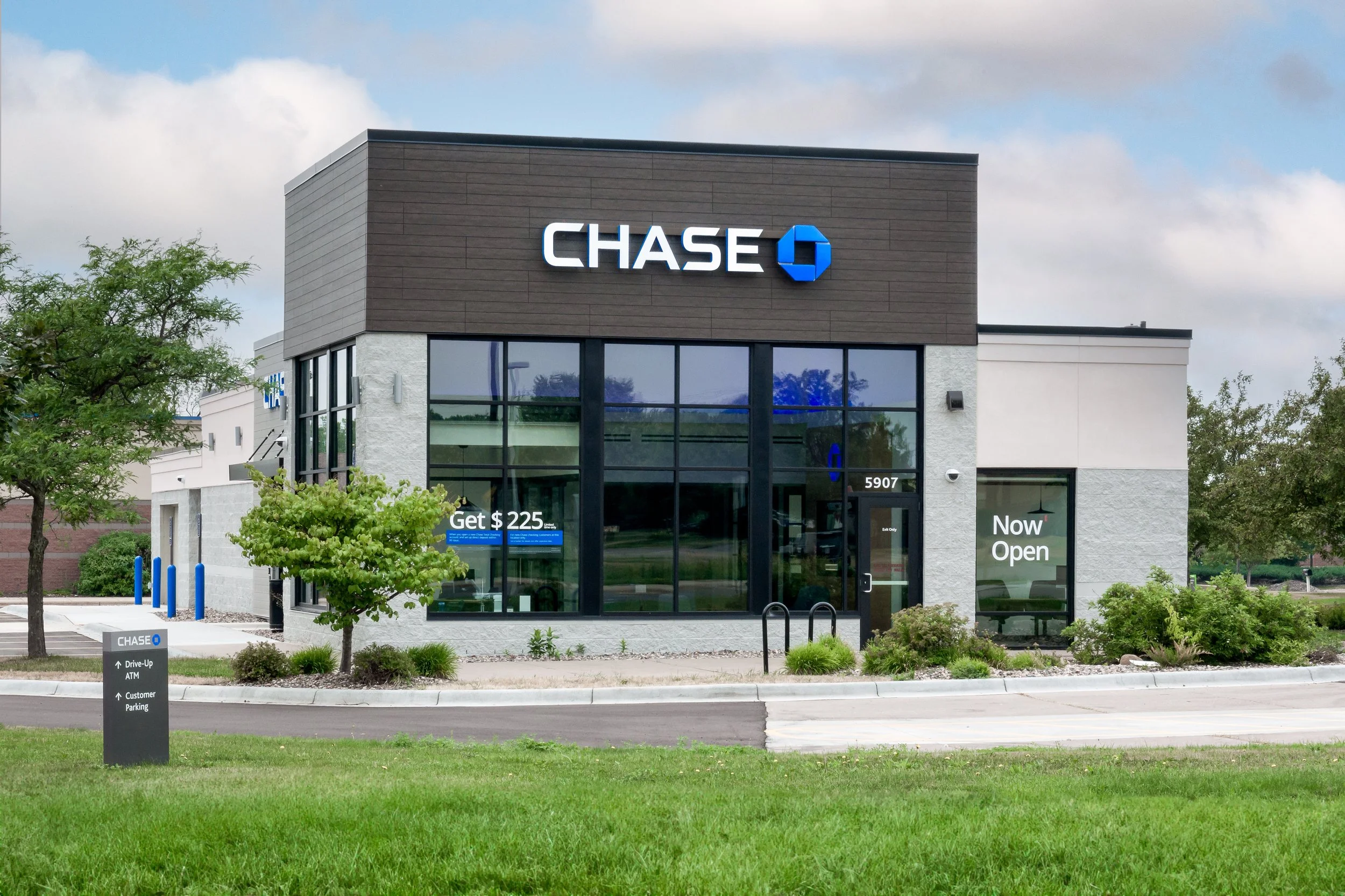 A Chase bank branch building with a sign on the front displaying the Chase logo and name, large front windows, and a small landscaped area with trees and bushes in front. Promotional signs are visible on the windows and sidewalk.