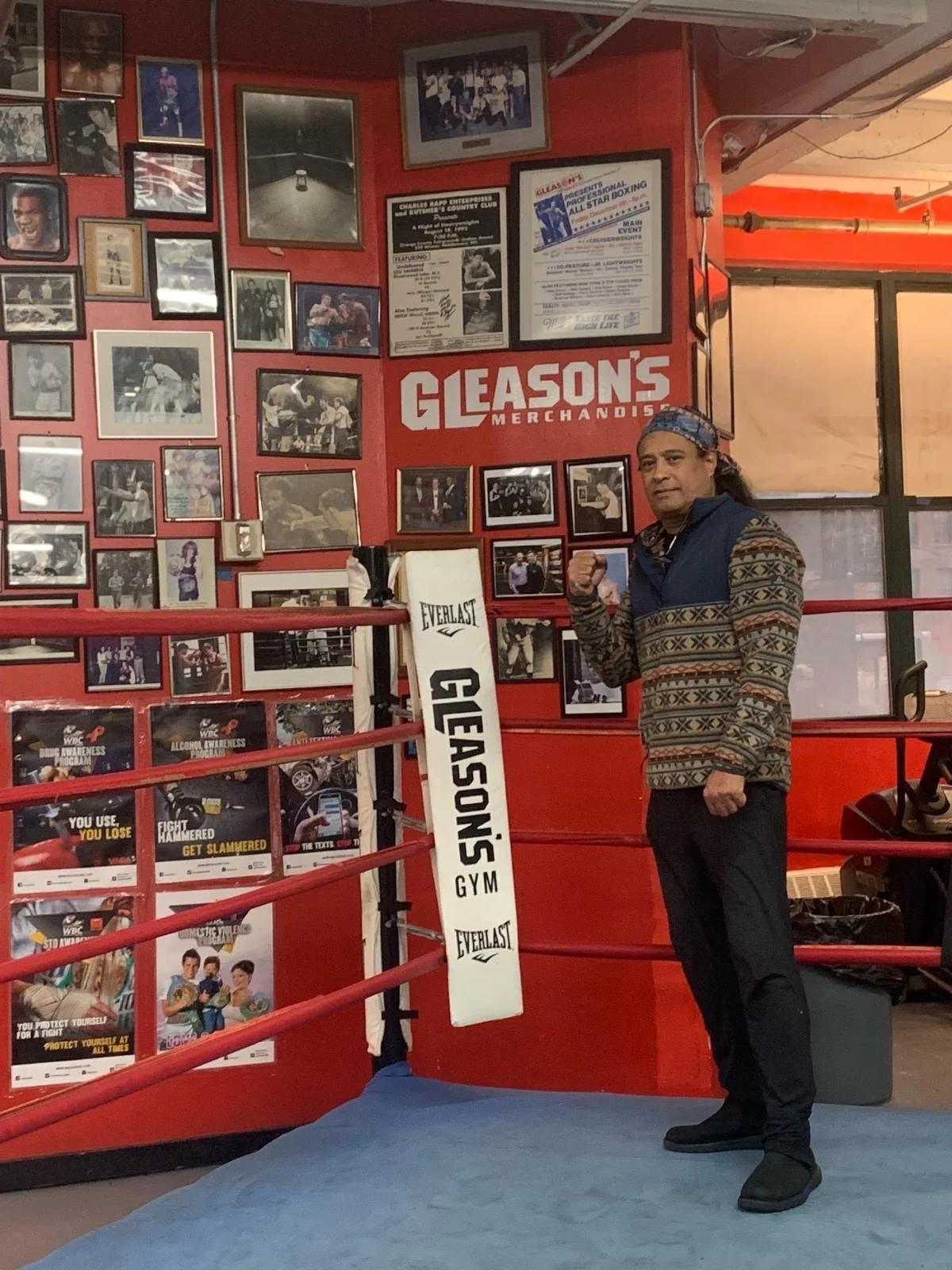 A man standing inside a boxing gym with red walls decorated with various framed photographs and posters, including one for Gleason's Merchandise. The man is wearing a patterned sweater, black pants, black shoes, and a colorful headband, and is making a fist gesture.