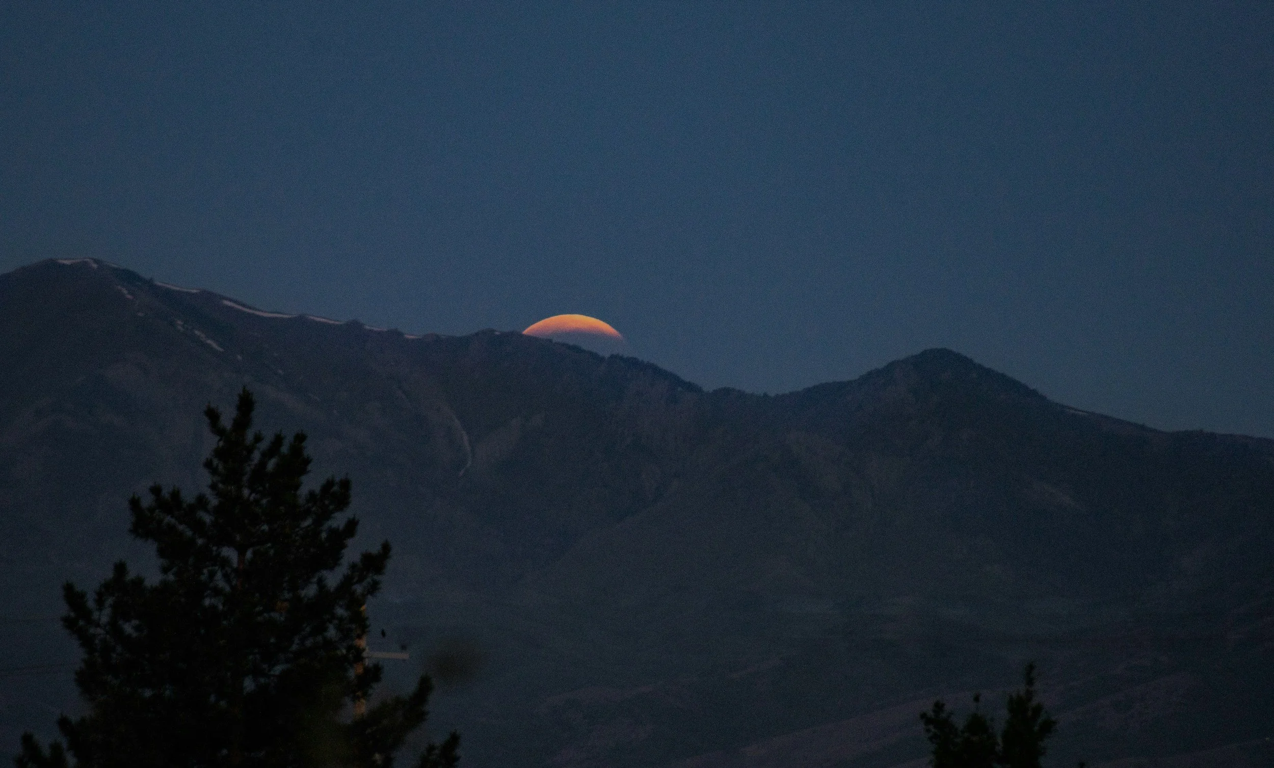 Night scene of mountains with a partly visible moon behind the peaks, dark blue sky, silhouettes of trees in the foreground.