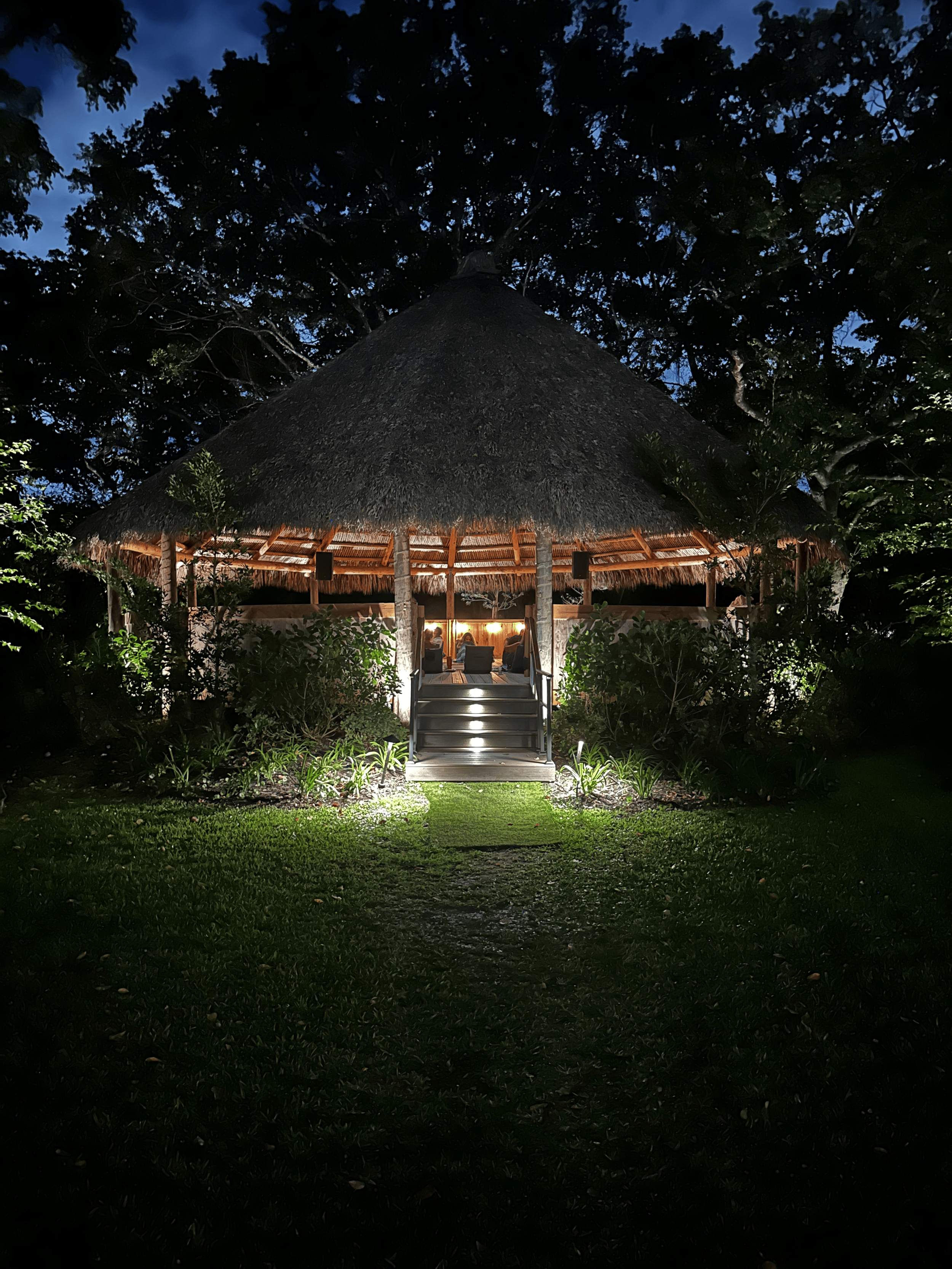 A thatched-roof hut with outdoor seating, illuminated at night, surrounded by greenery and trees.