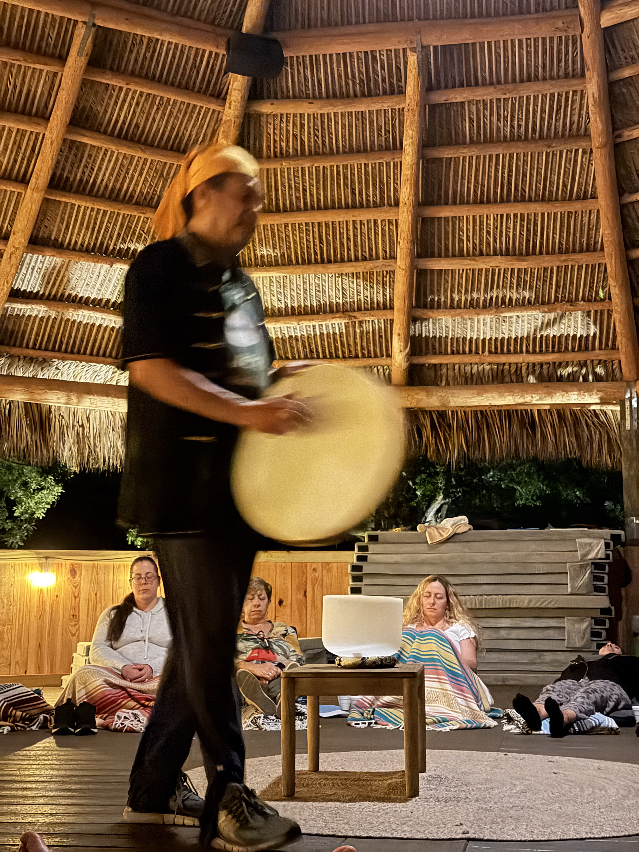 People sitting on a floor under a thatched roof, some with eyes closed, others lying down, with a woman standing and holding a large object in a dimly lit room.