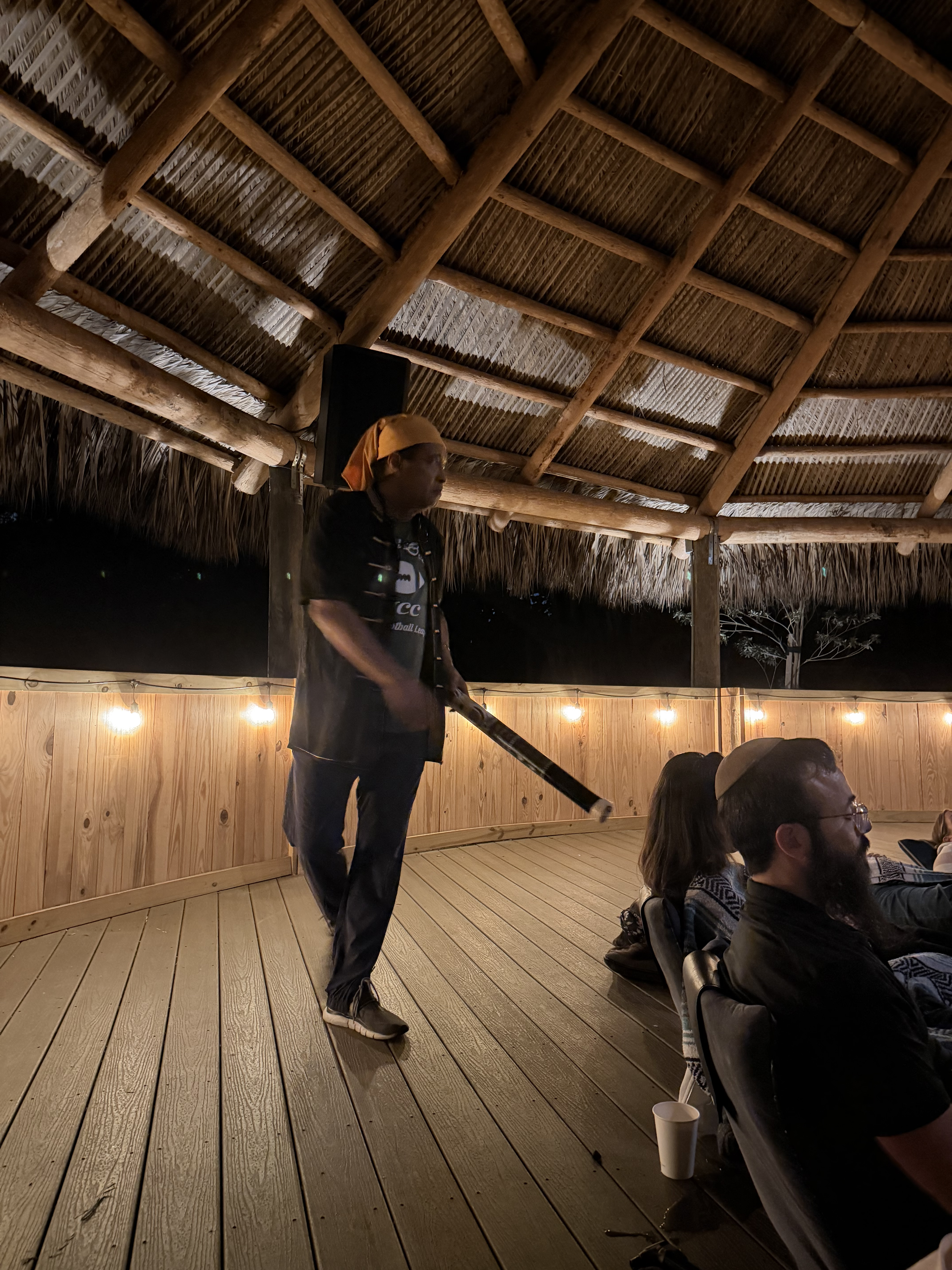A person wearing a yellow head wrap and black attire is playing a musical instrument, possibly a flute or similar, on a wooden deck under a thatched roof with string lights. There are seated people watching, and a black speaker is mounted on a post.