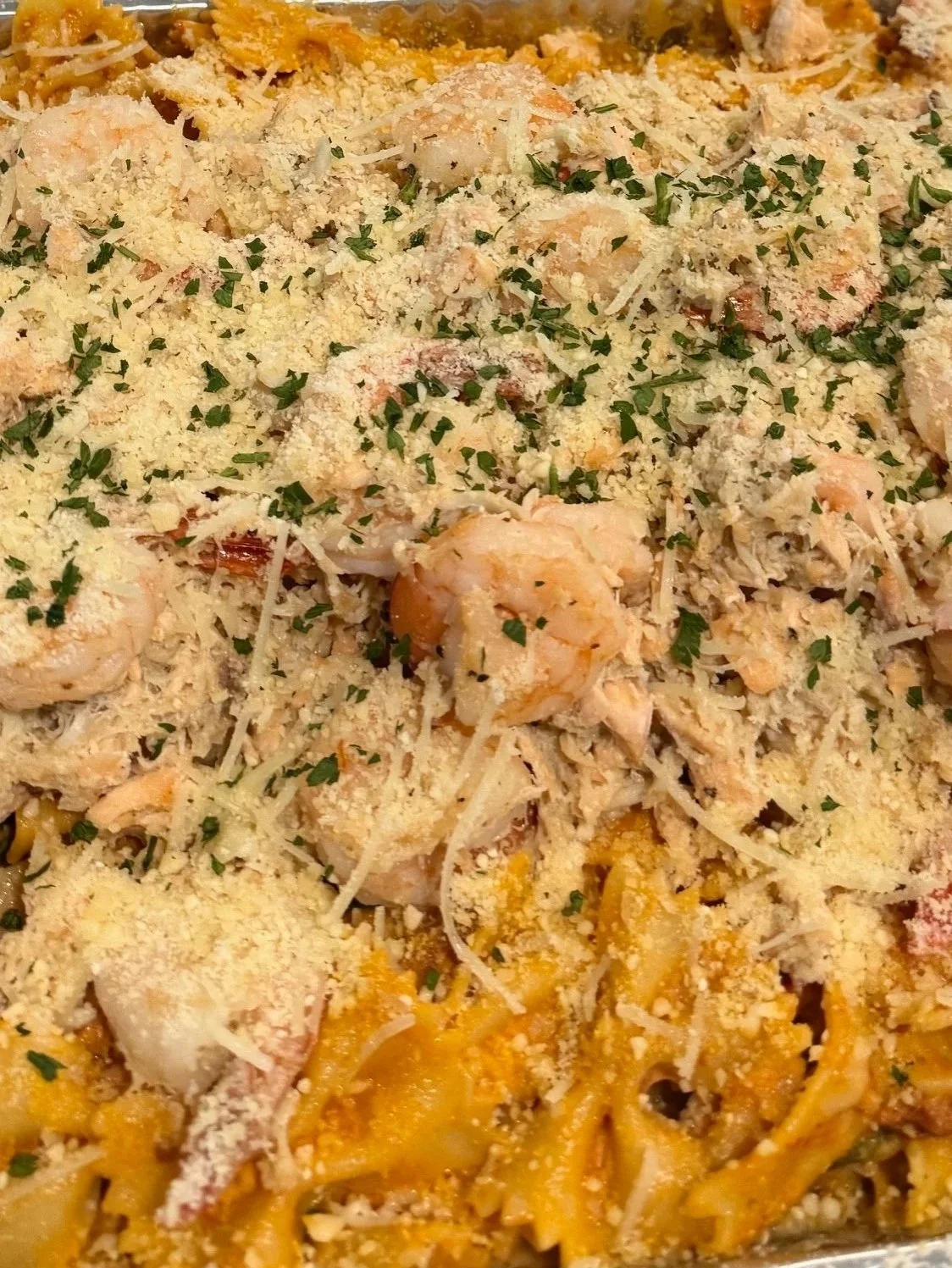 Close-up of a tray of baked pasta topped with shredded cheese, chopped herbs, and breadcrumbs.