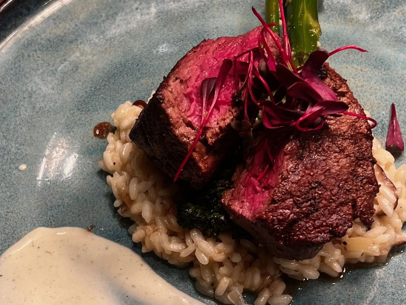 A plated dish featuring medium-rare beef tenderloin slices on a bed of creamy risotto, garnished with fresh microgreens and accompanied by a dollop of creamy sauce.