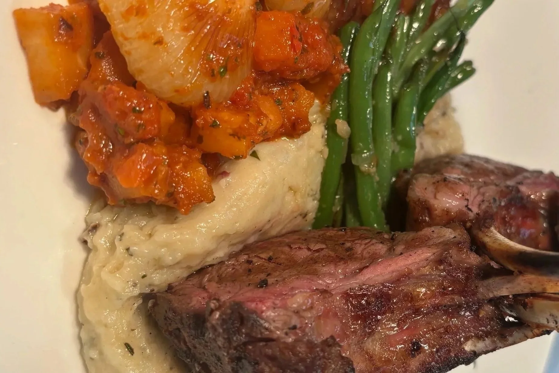 Close-up of a plated meal featuring cooked steak, mashed potatoes topped with a spoonful of chunky vegetable stew, green beans, and a slice of roasted sweet potato.