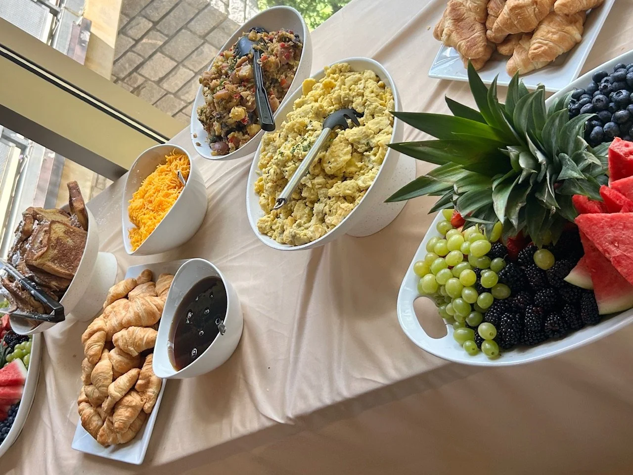 Assorted breakfast foods on a table, including scrambled eggs, potato salad, croissants, flaky rolls, fresh fruit like watermelon, blackberries, green and red grapes, pineapples, and blueberries.