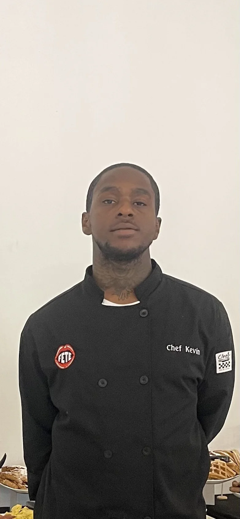 Portrait of a man wearing a black chef coat with patches and embroidery, standing indoors in front of a plain white wall.