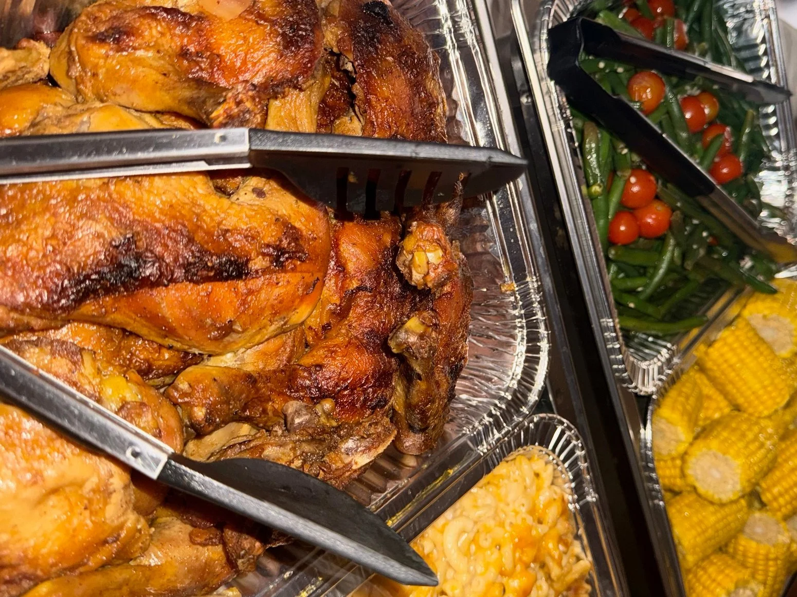 Close-up view of a buffet with roasted chicken, green beans with cherry tomatoes, corn on the cob, and macaroni and cheese.