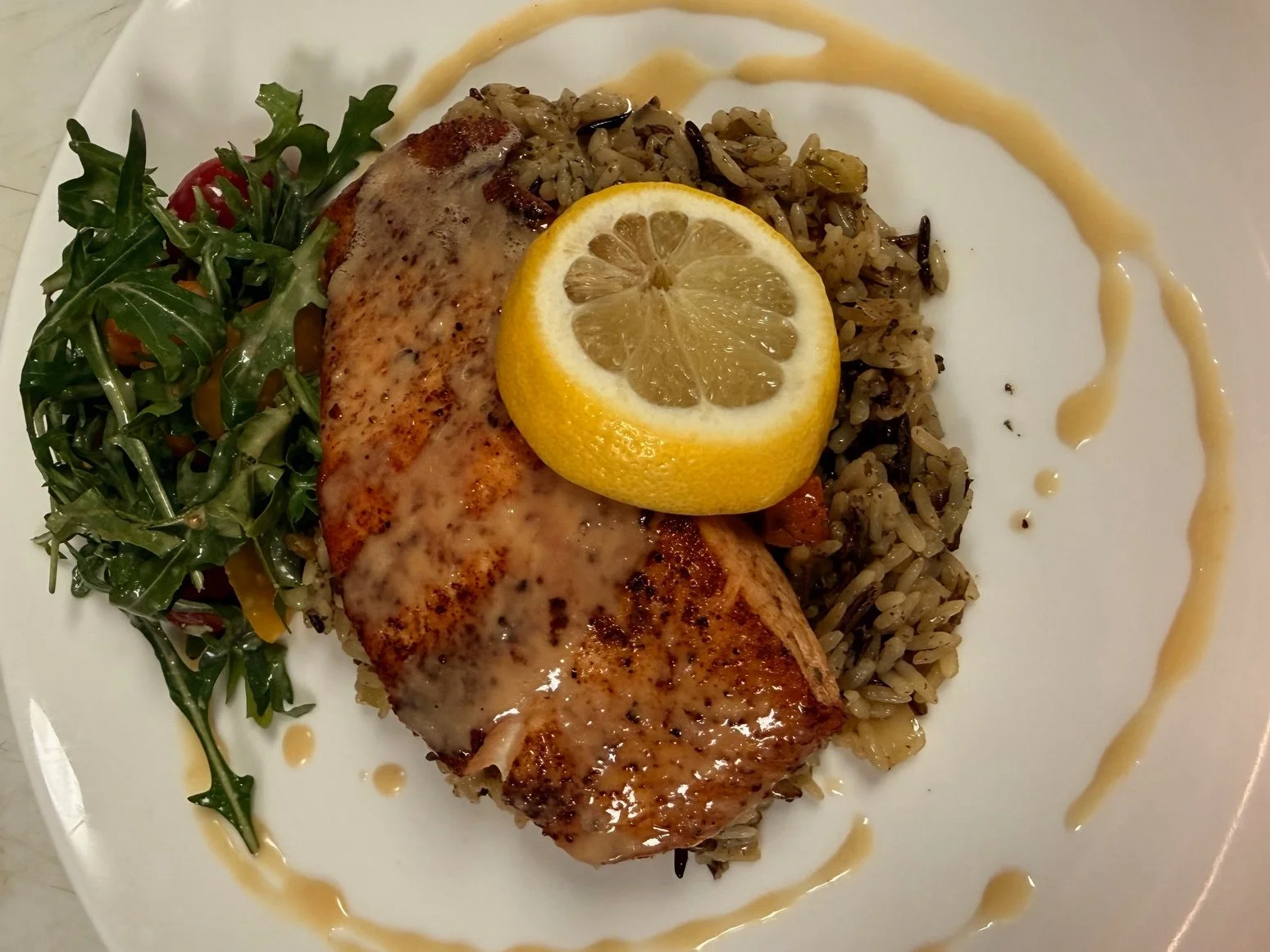 A plate with grilled salmon topped with a lemon slice, served with wild rice, a side salad of greens, and a drizzle of sauce.