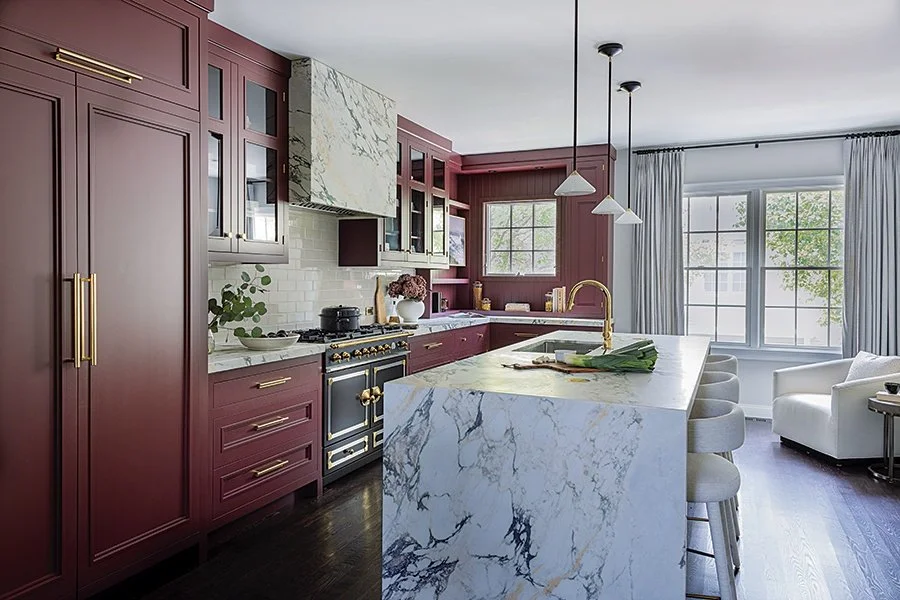 Modern kitchen with burgundy cabinets, marble island, stainless steel appliances, gold accents, and large windows with white curtains.