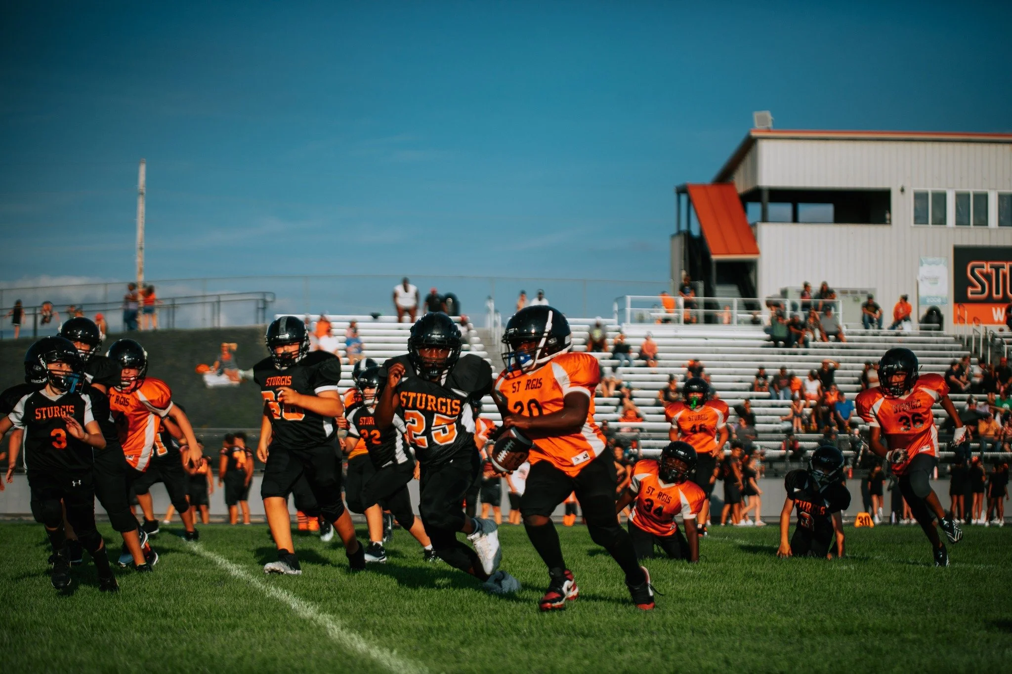 American football players in orange and black uniforms running on a football field with spectators in the stands and a building in the background.