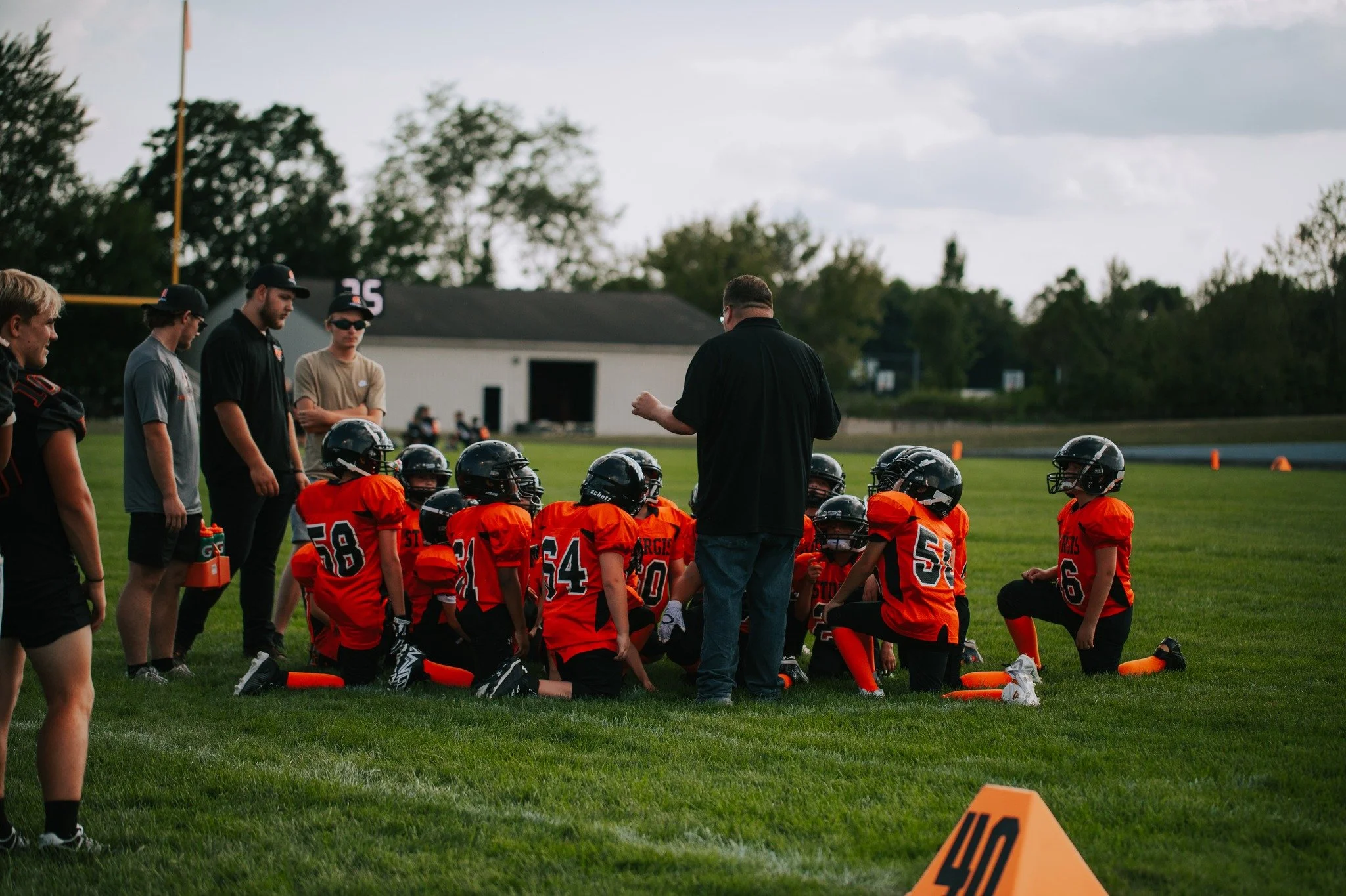 A youth football team gathered on a field with their coach, listening attentively during a practice or game. The players are dressed in orange uniforms with black helmets, and a few adults, possibly coaches or parents, are standing nearby. Orange cones and yardage markers are visible on the grass playing field, with a building and trees in the background.