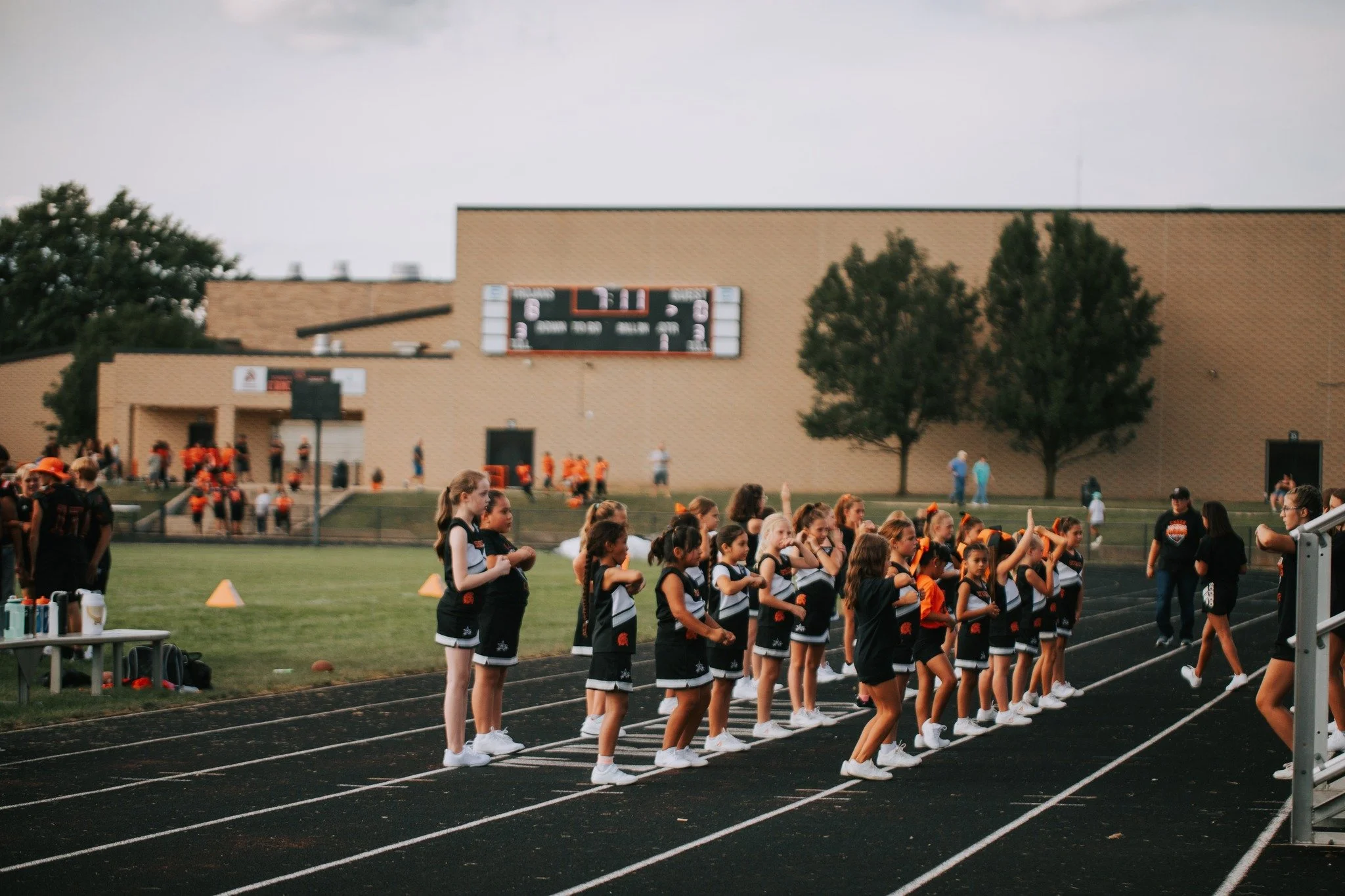 Group of young cheerleaders standing in a line on a running track during a sports event, with a scoreboard and spectators in the background.