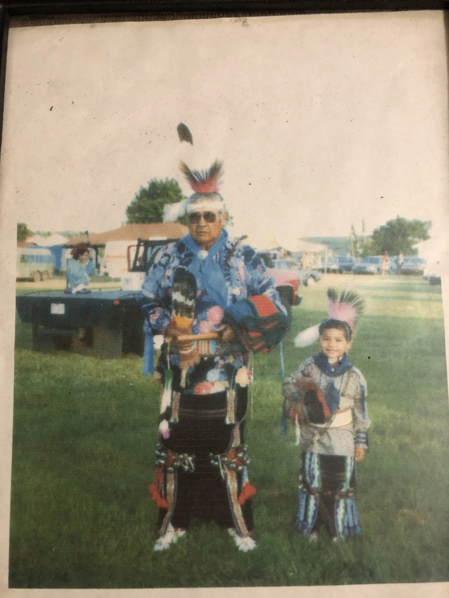 A man and a young boy dressed in traditional Native American attire standing outdoors on grass, with a celebration or festival in the background. The man is wearing sunglasses, a blue shirt with a floral pattern, and a feathered headdress. The boy is dressed in a matching traditional outfit and also wears a feathered headdress.