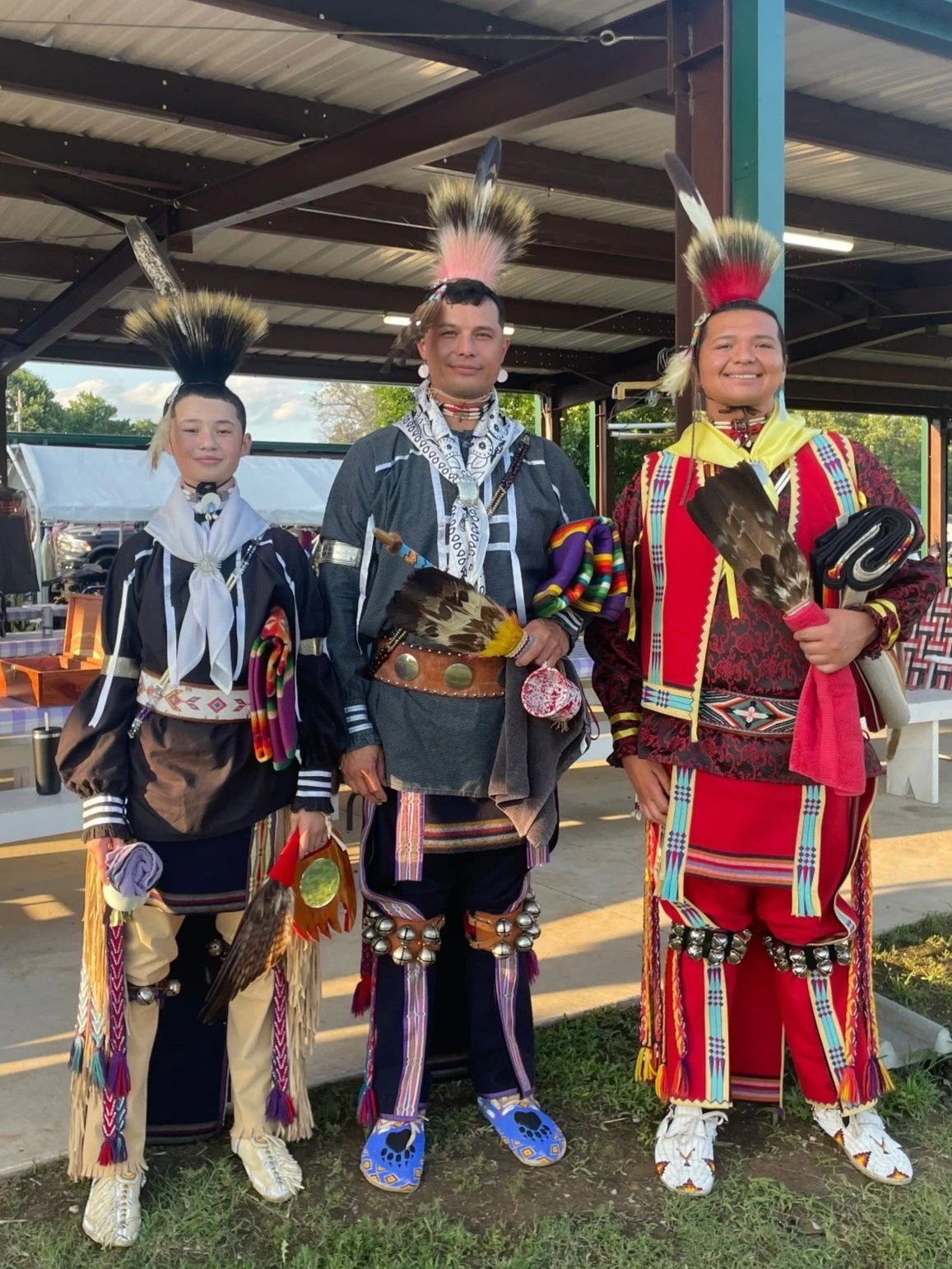 Three individuals dressed in traditional Native American attire, standing outdoors under a metal pavilion, with one person holding a fan made of feathers.