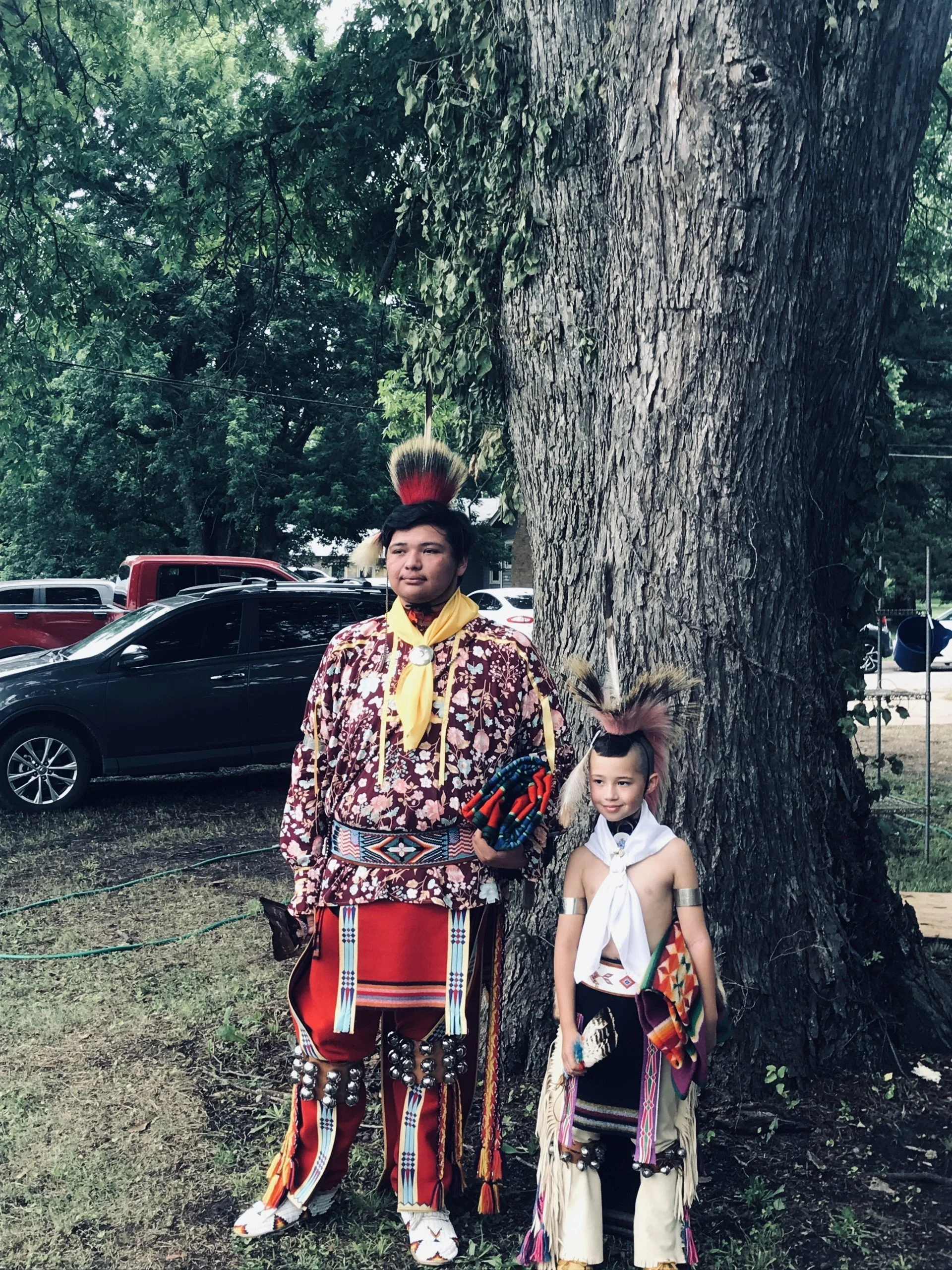 Two children dressed in traditional indigenous clothing standing outdoors near a large tree, with parked cars in the background.