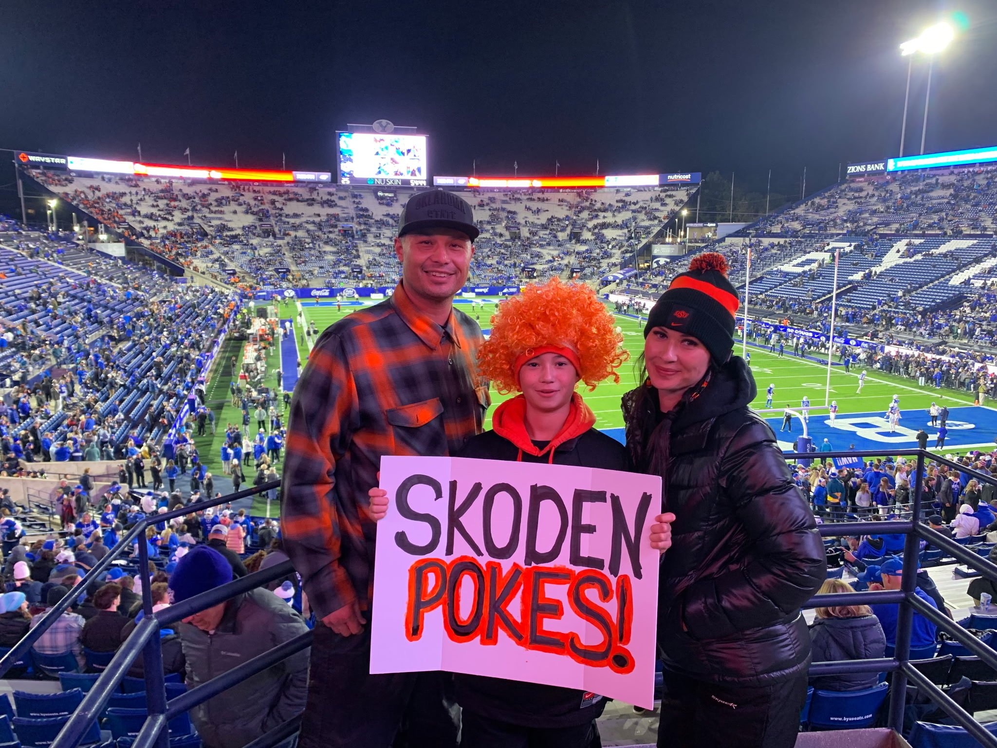 Three people standing in a football stadium at night, holding a sign that reads 'Skoden Pokes!' The man on the left is wearing a dark baseball cap and a plaid shirt, the girl in the middle is wearing an orange curly wig and an orange hoodie, and the woman on the right is wearing a black winter hat with orange and black pom-poms and a black jacket. The stadium is filled with people and the field is visible in the background.