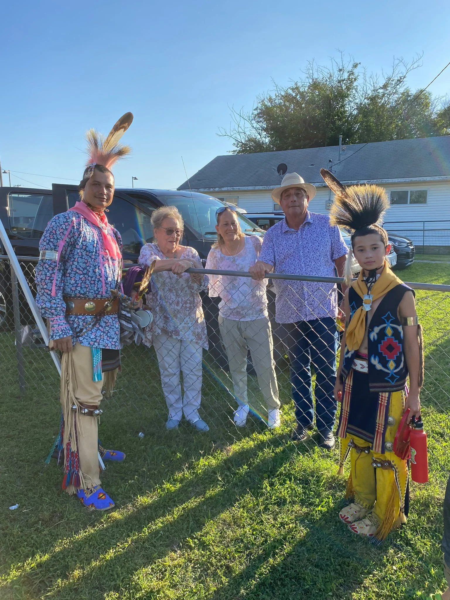 A group of six people standing outside behind a chain-link fence, with three dressed in Native American regalia and three in casual clothes. The scene is sunny with a blue sky, and there are cars and a house in the background.