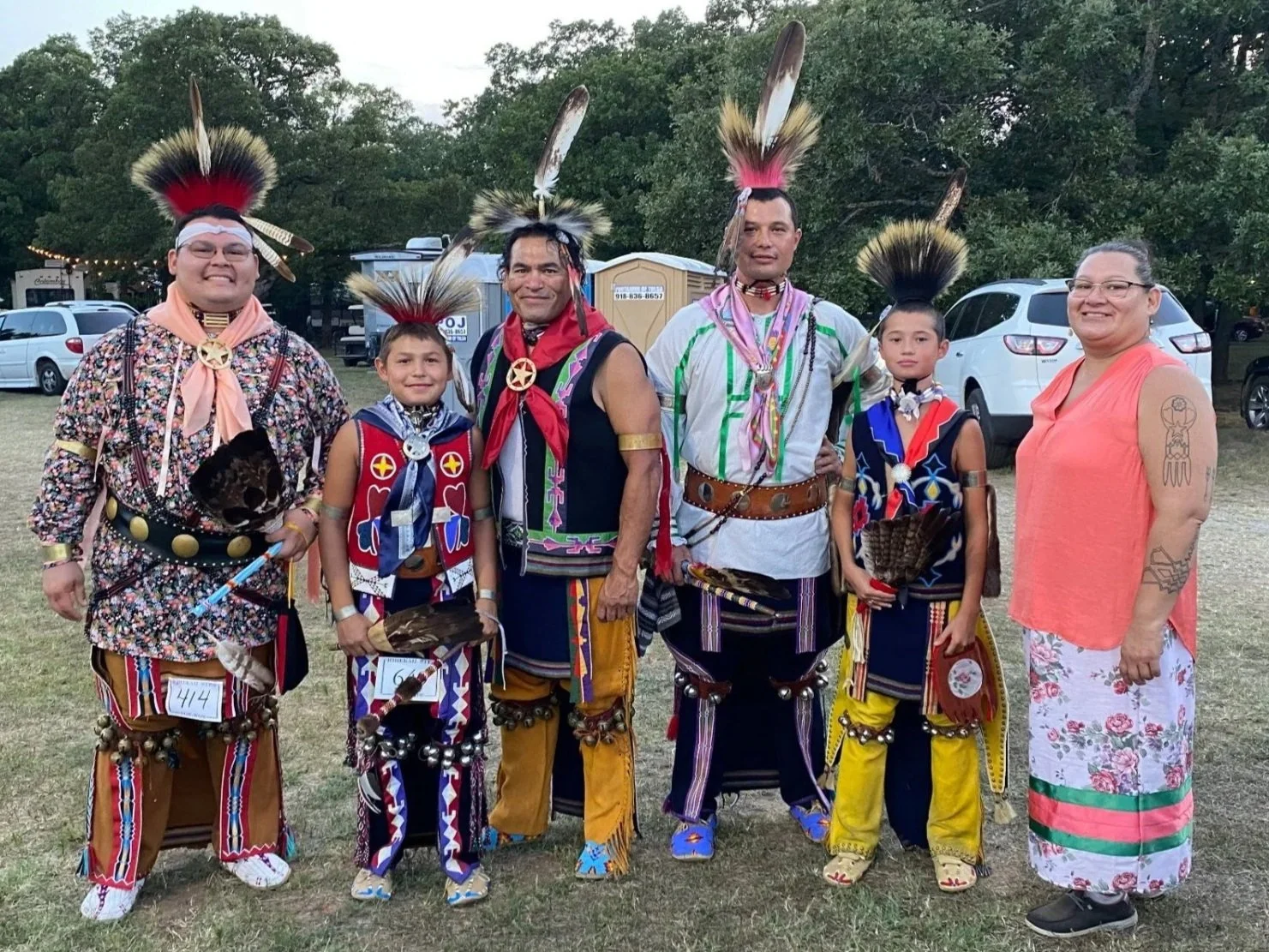Group of people dressed in traditional Native American clothing, including feathered headdresses, beadwork, and colorful attire, outdoors at a gathering or event.