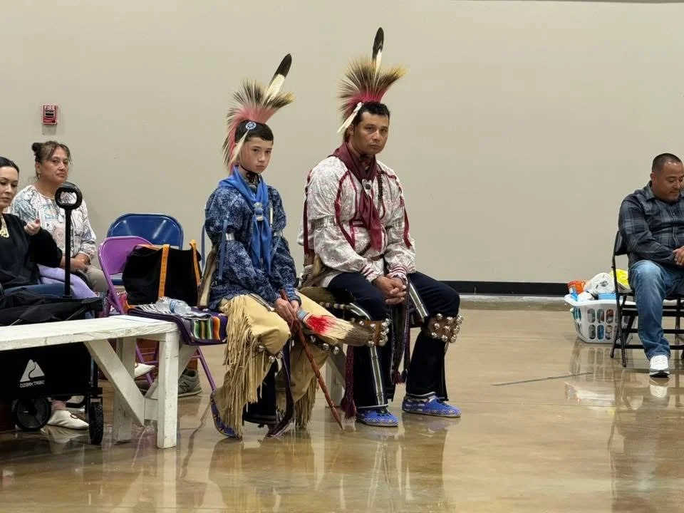 Two Native American men dressed in traditional regalia with feather headdresses sitting on chairs in the center, surrounded by seated women and a man in casual clothing, inside a community room.