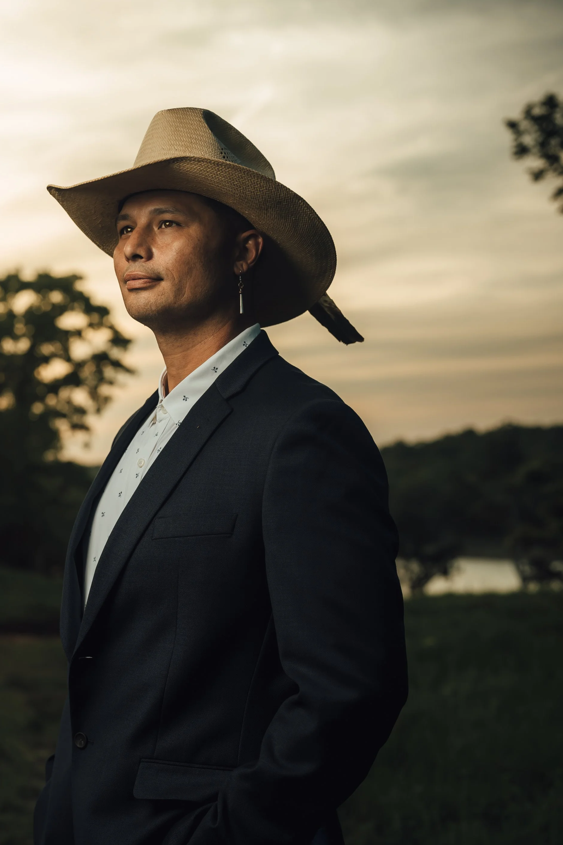 A man in a suit with a large straw hat standing outdoors during sunset with trees and a body of water in the background.