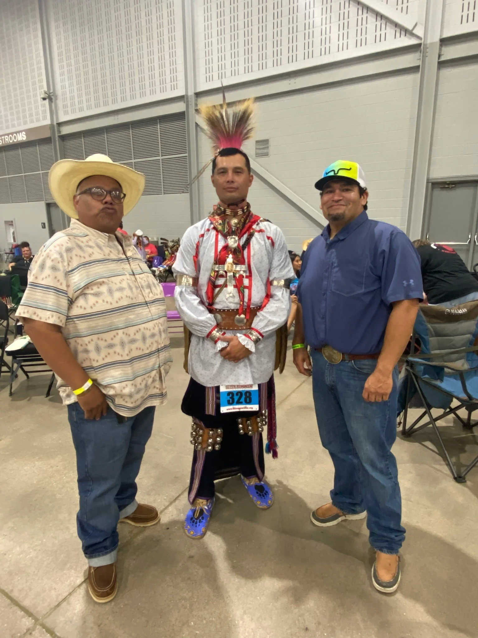 Three men standing indoors, one dressed in traditional Native American attire and the other two in casual Western-style clothing.